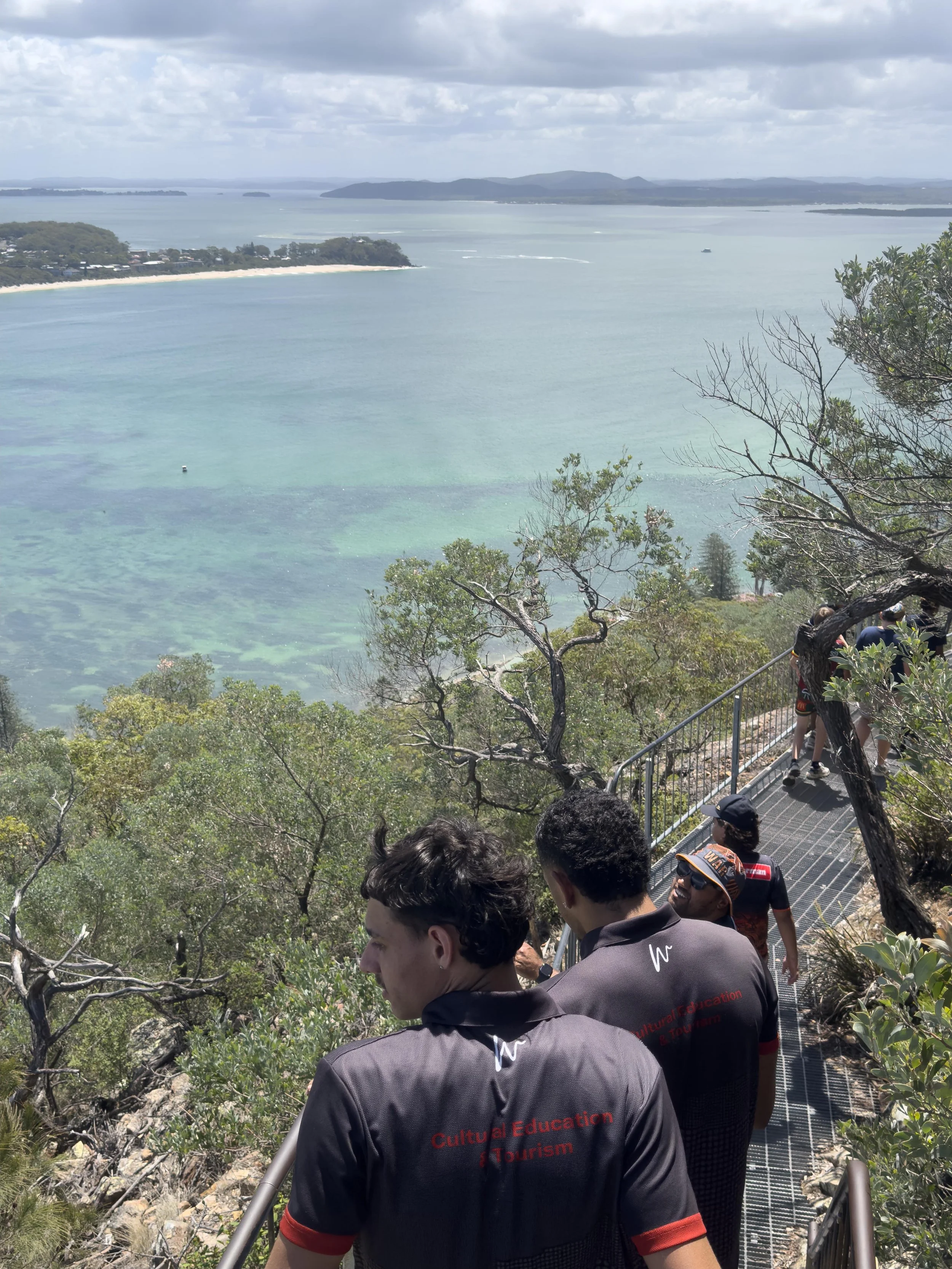 People walking down a walkway with trees, overlooking a large body of water with islands and a distant shoreline under cloudy sky.