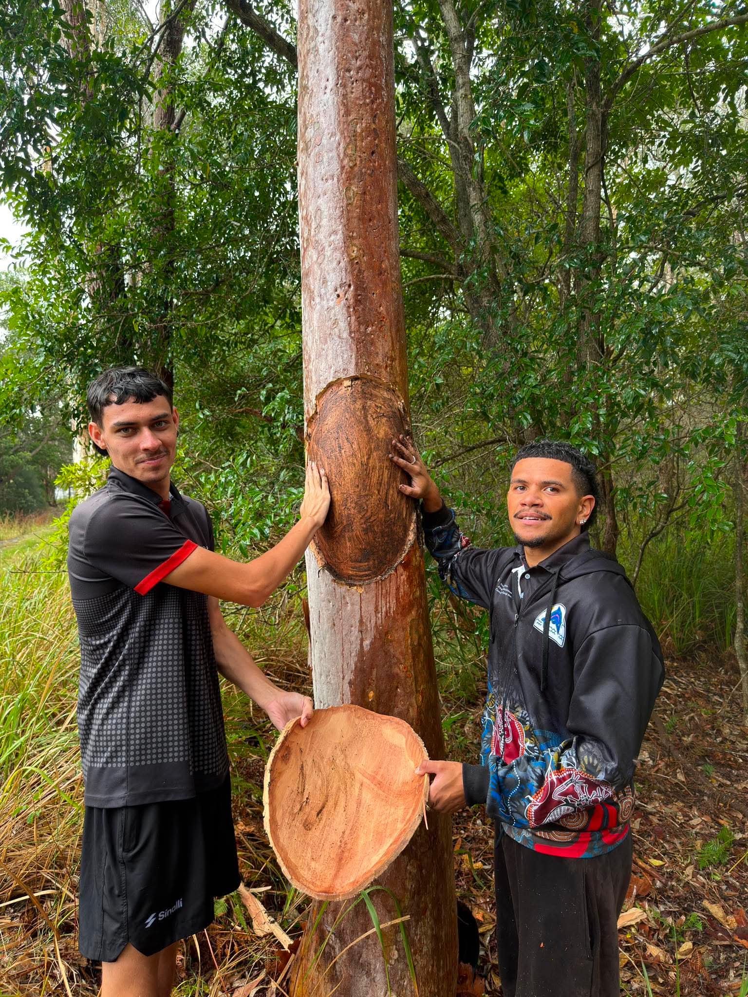 Two men are standing outdoors in a forested area, holding a large tree trunk. They are posing for the photo, with greenery in the background.