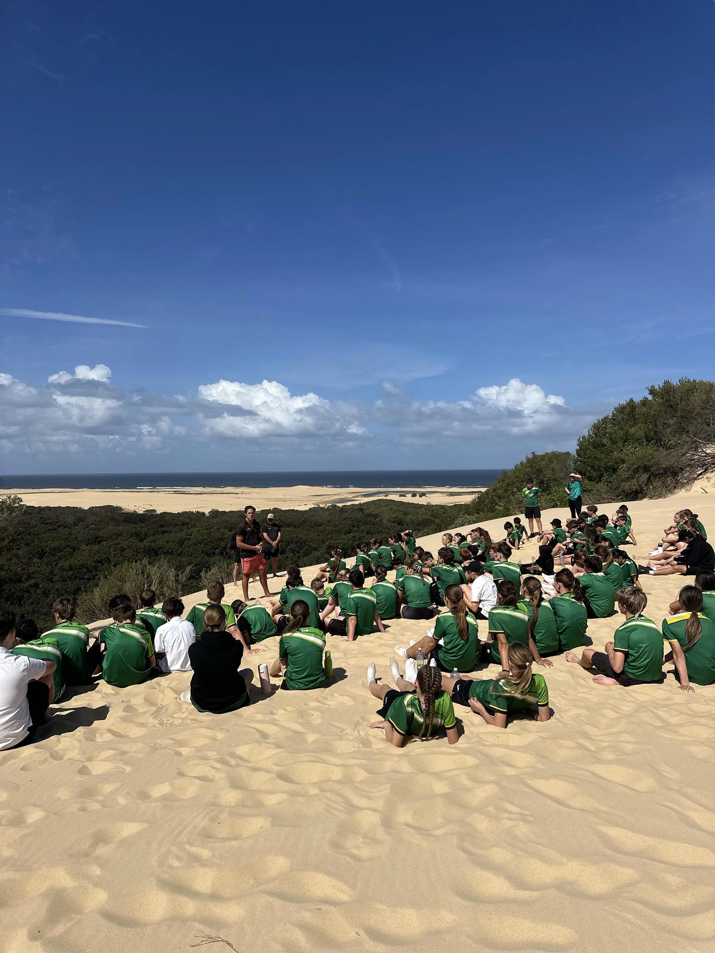 A large group of students sitting on sandy dunes.