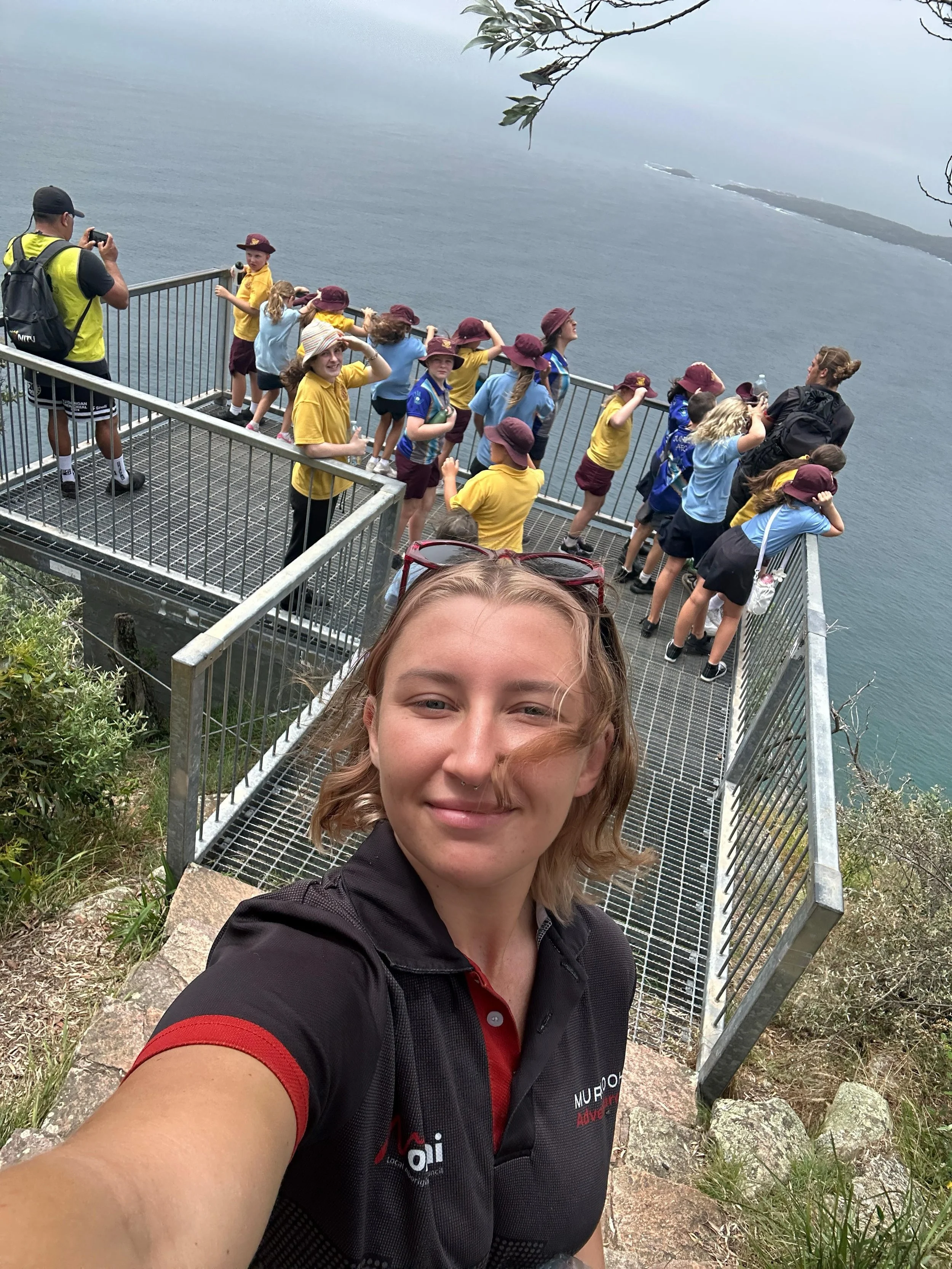 A young woman taking a selfie on a scenic overlook with a group of children in school uniforms and hats.
