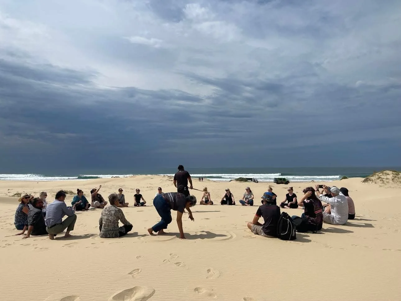 A group of people sitting in a circle on a sandy beach with the ocean and cloudy sky in the background. Some people are taking photos, and two individuals are standing or squatting in the center of the circle.