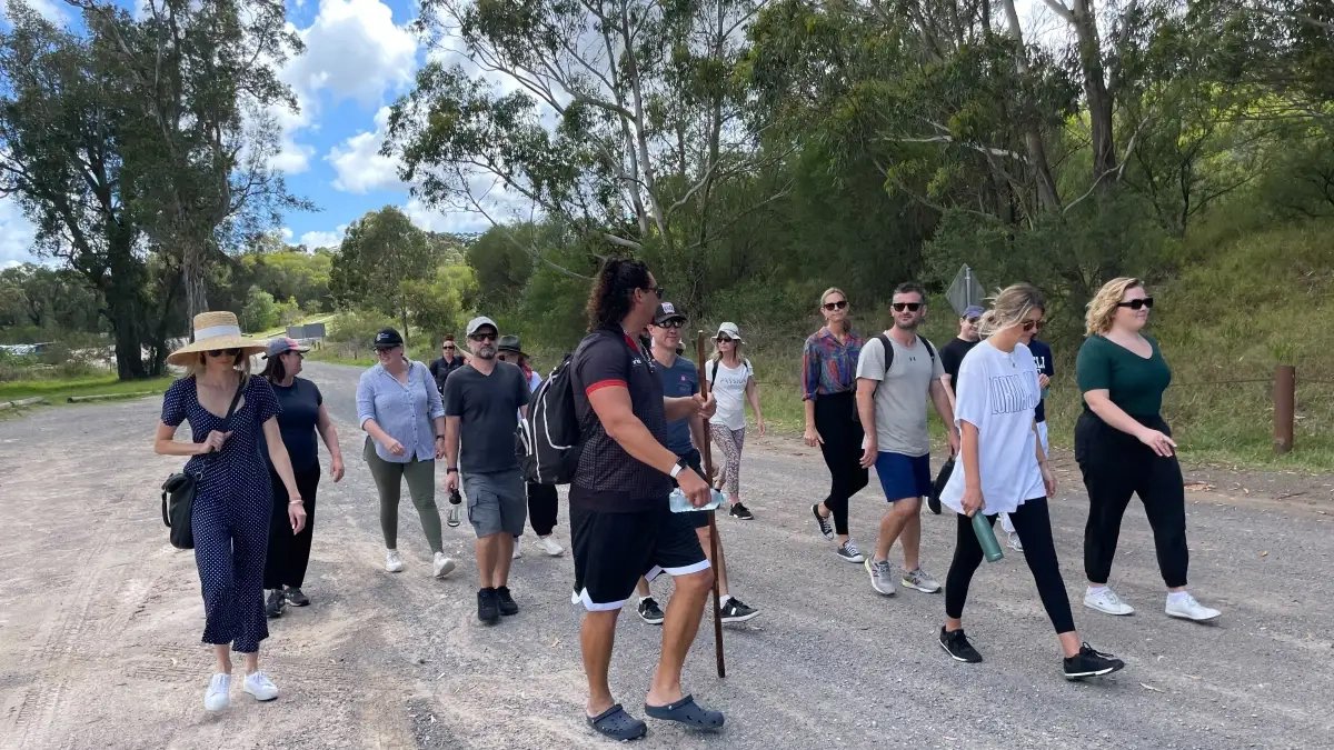 A group of people walking outdoors on a gravel path surrounded by trees and greenery, on a partly cloudy day.
