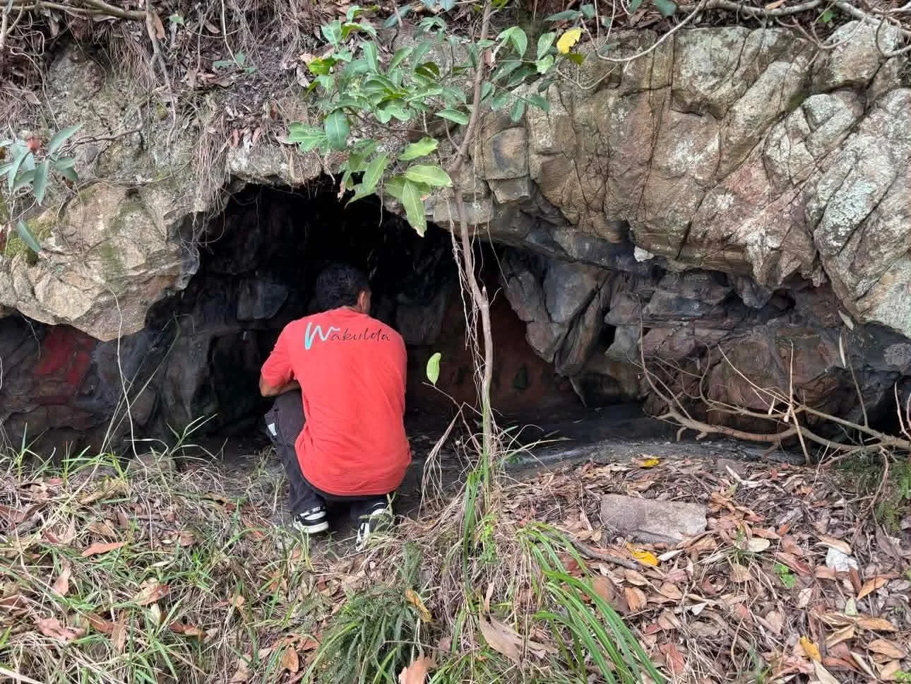 Person crouching in front of a cave entrance surrounded by rocks and dry leaves.