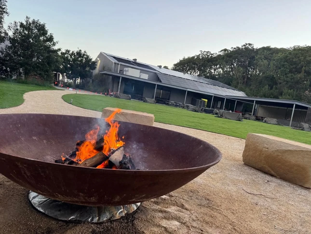 A fire pit with burning logs in front of a modern building, surrounded by a gravel pathway, grass, and trees.