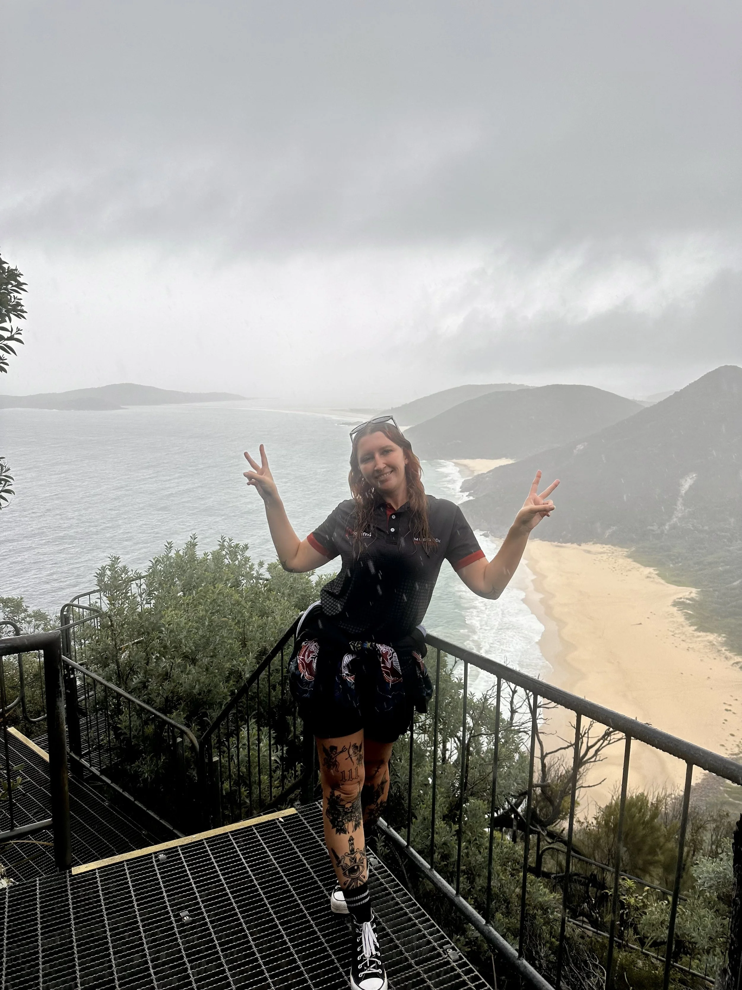 A woman stands on a metal viewing platform overlooking a foggy beach with cliffs and ocean in the background. She is making peace signs with both hands, smiling, and wearing a dark sports shirt, patterned leggings, and sneakers.