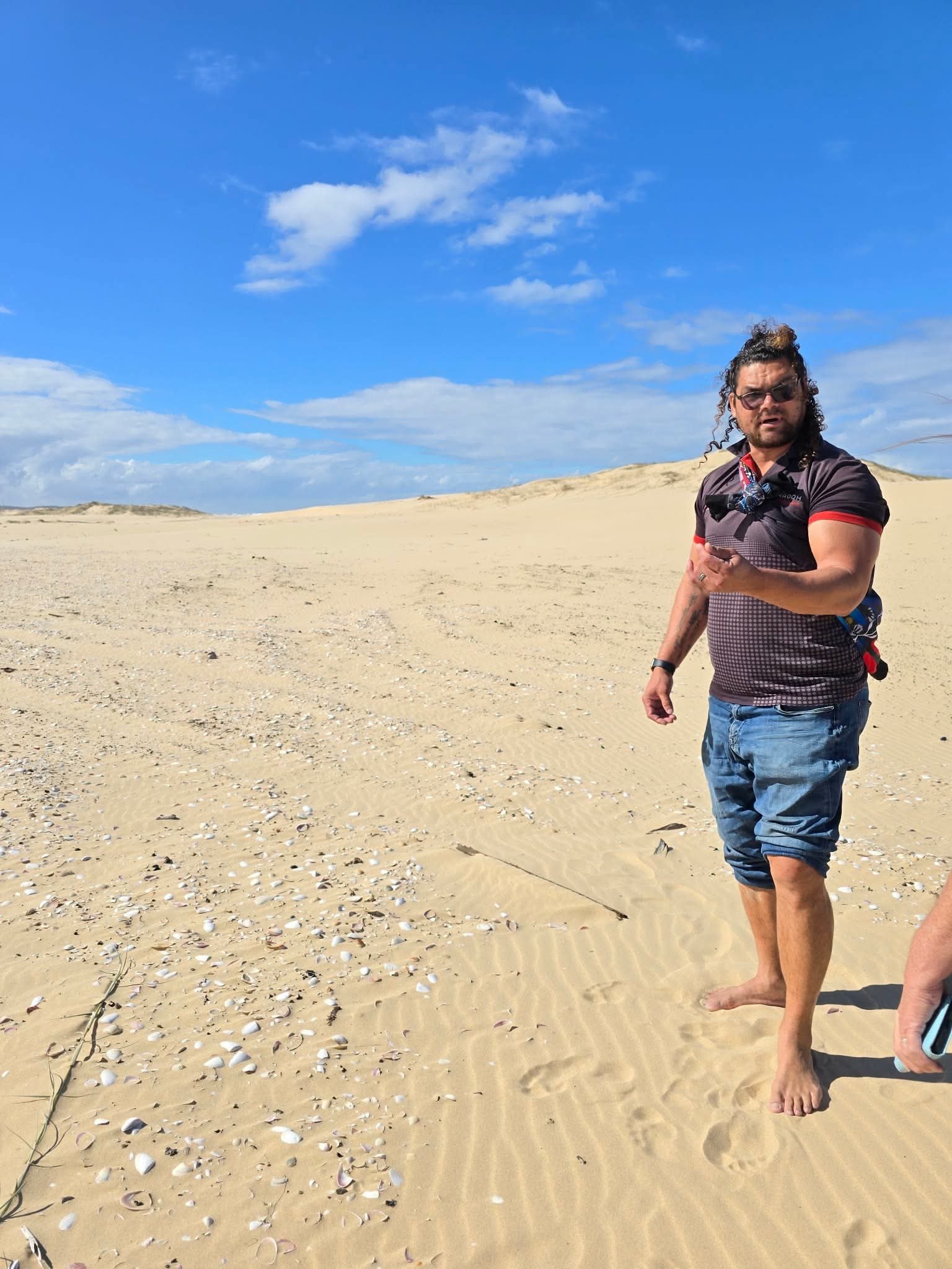 A man with curly hair, sunglasses, wearing a black and gray athletic shirt and blue shorts, walking barefoot on a sandy beach with shells, with sand dunes in the background and a bright blue sky with scattered clouds.