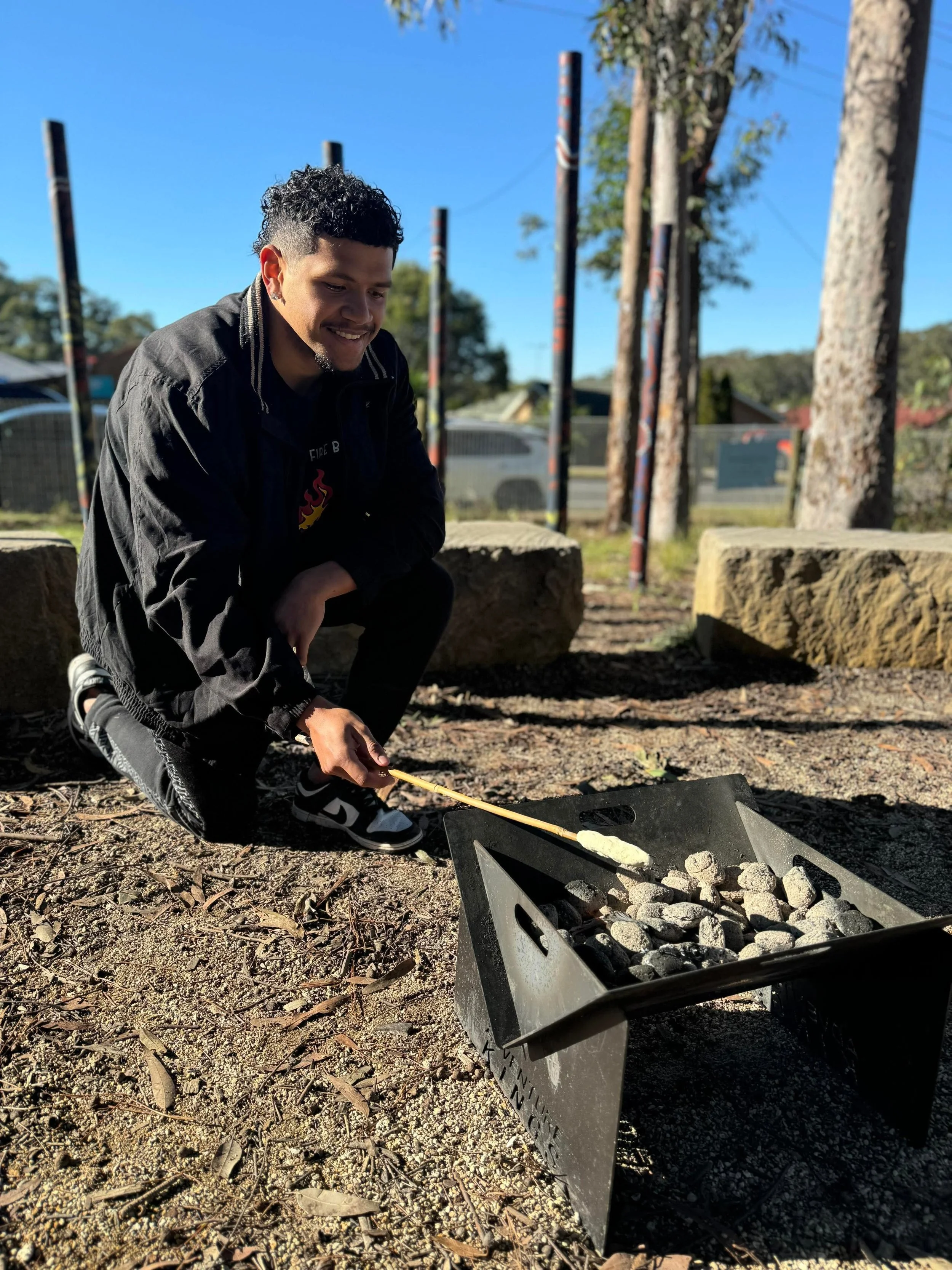 A young man with curly hair, wearing a black jacket and sneakers, is crouching and painting a rock white outdoors. He is smiling and appears to be enjoying the activity with a clear blue sky in the background.