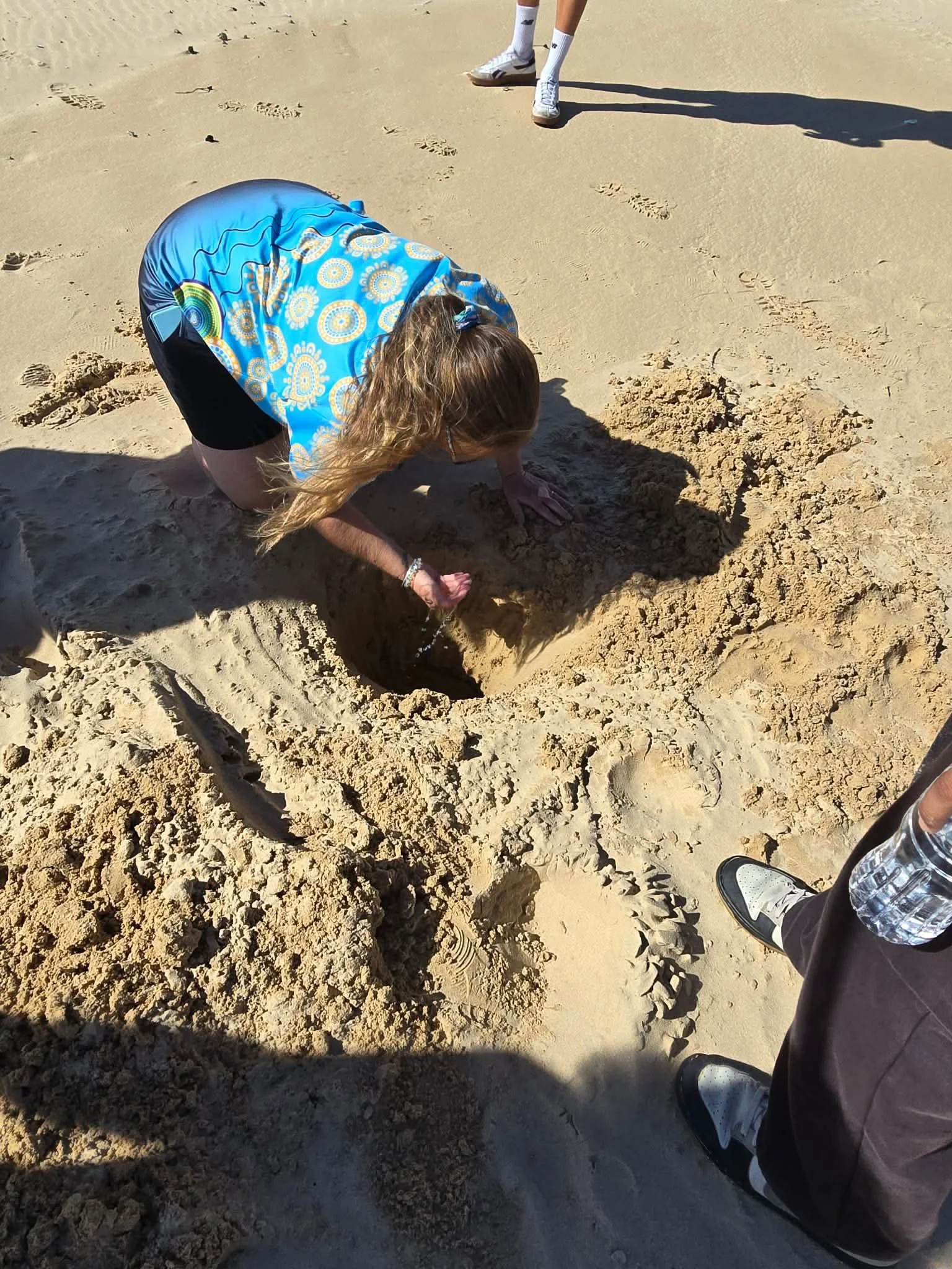 A person is kneeling on sandy ground, digging a hole with their hands. They are wearing a blue patterned shirt, black shorts, and have long, light brown hair. There are footprints and marks on the sand around them.