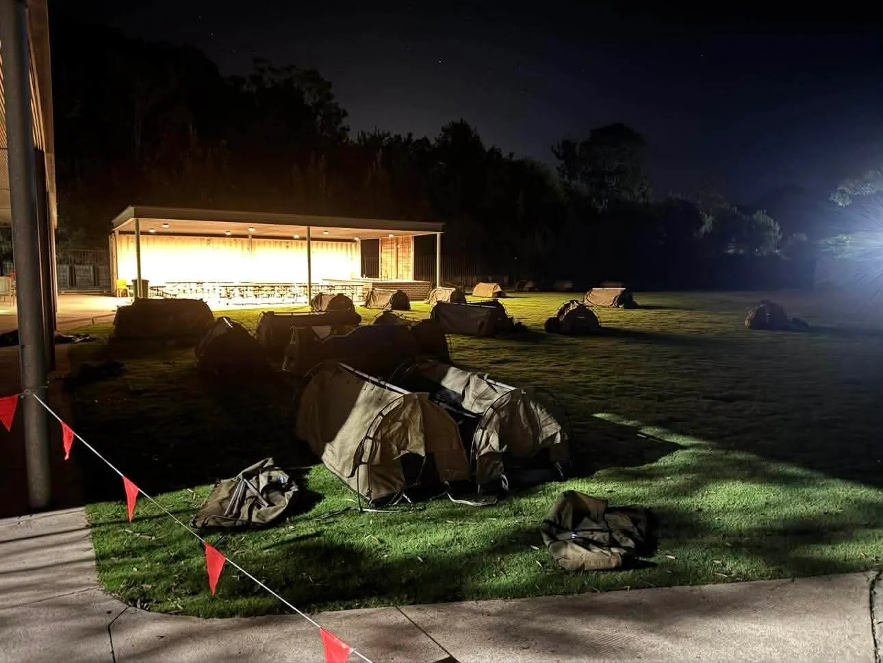 Nighttime scene of tents set up on a grassy field with a lit pavilion structure in the background and trees behind it, illuminated by bright outdoor lights.
