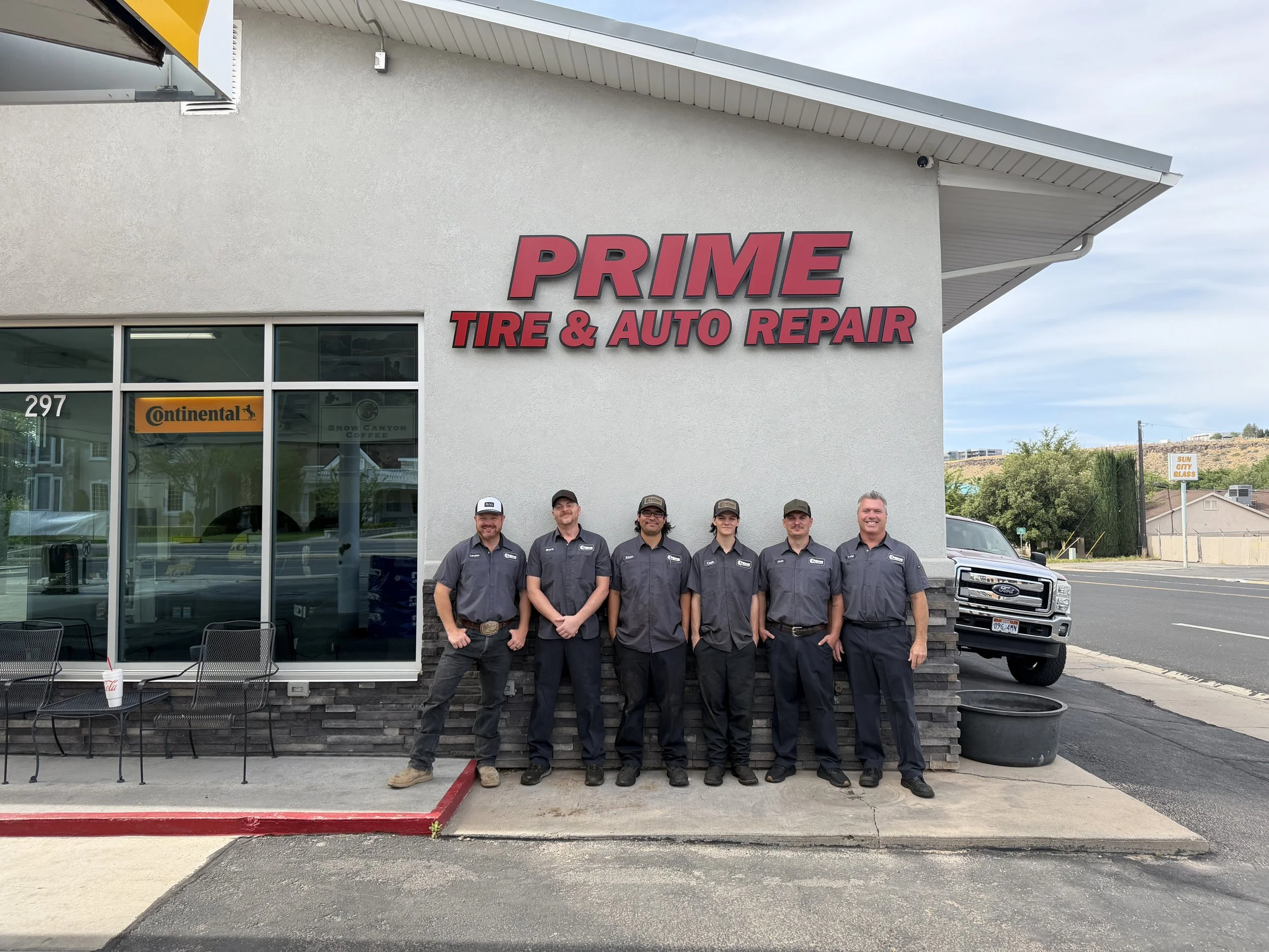 Group of six auto repair mechanics standing in front of a tire and auto repair shop named 'PRIME TIRE & AUTO REPAIR,' with storefront windows reflecting the St. George Blvd., ready to answer the needs of customers that search "auto repair near me."