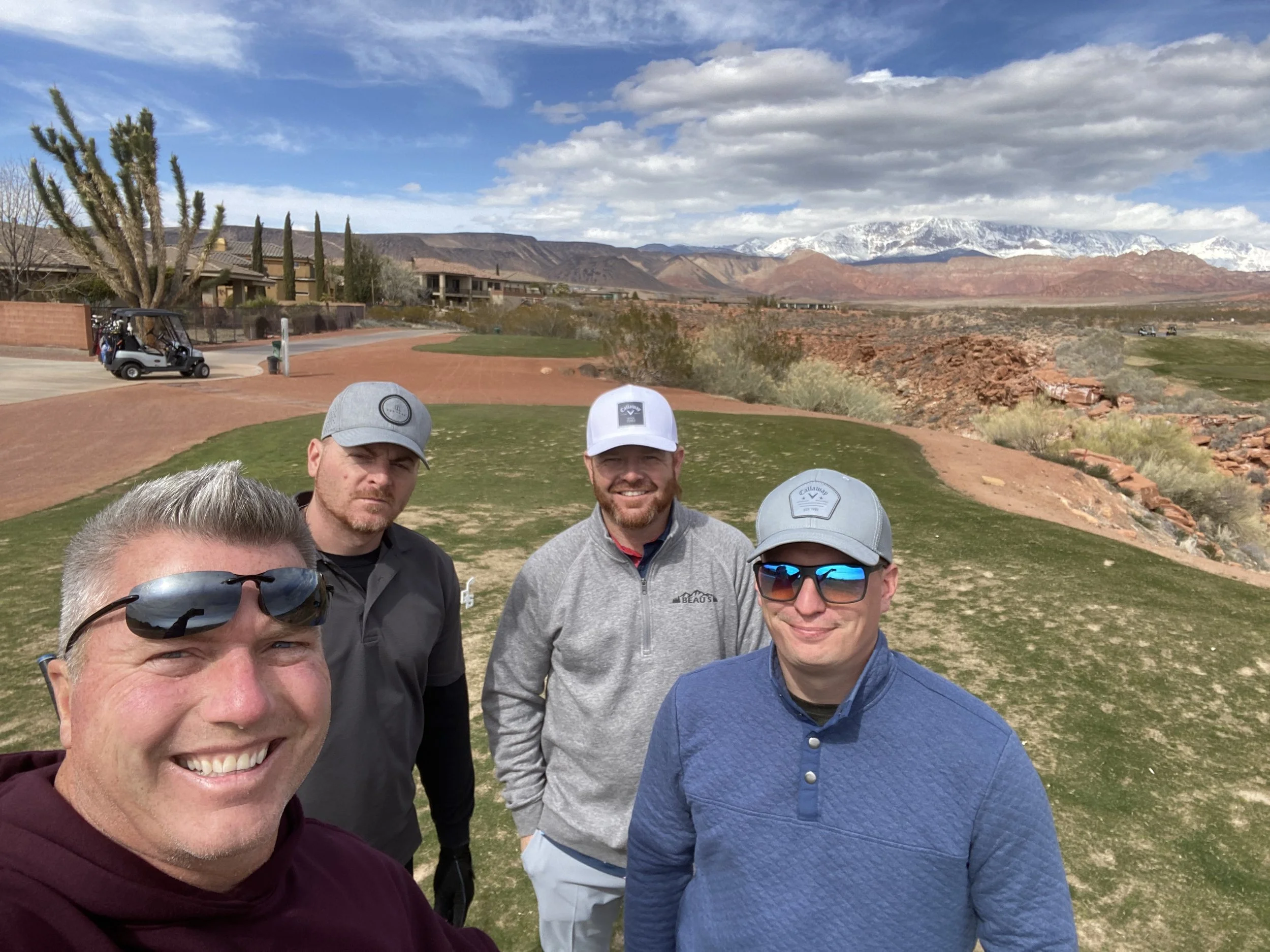 Group of four men from Prime Tire and Auto Repair taking a selfie on a St. George golf course with mountains in the background. Snow-capped peaks, a blue sky with clouds, and dry desert terrain visible.