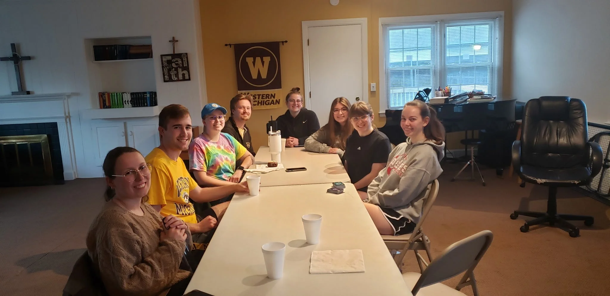 A group of nine young people sitting around a long table in a room, smiling at the camera. The room has a window showing a building outside, a West Michigan banner on the wall, and various office items and furniture.
