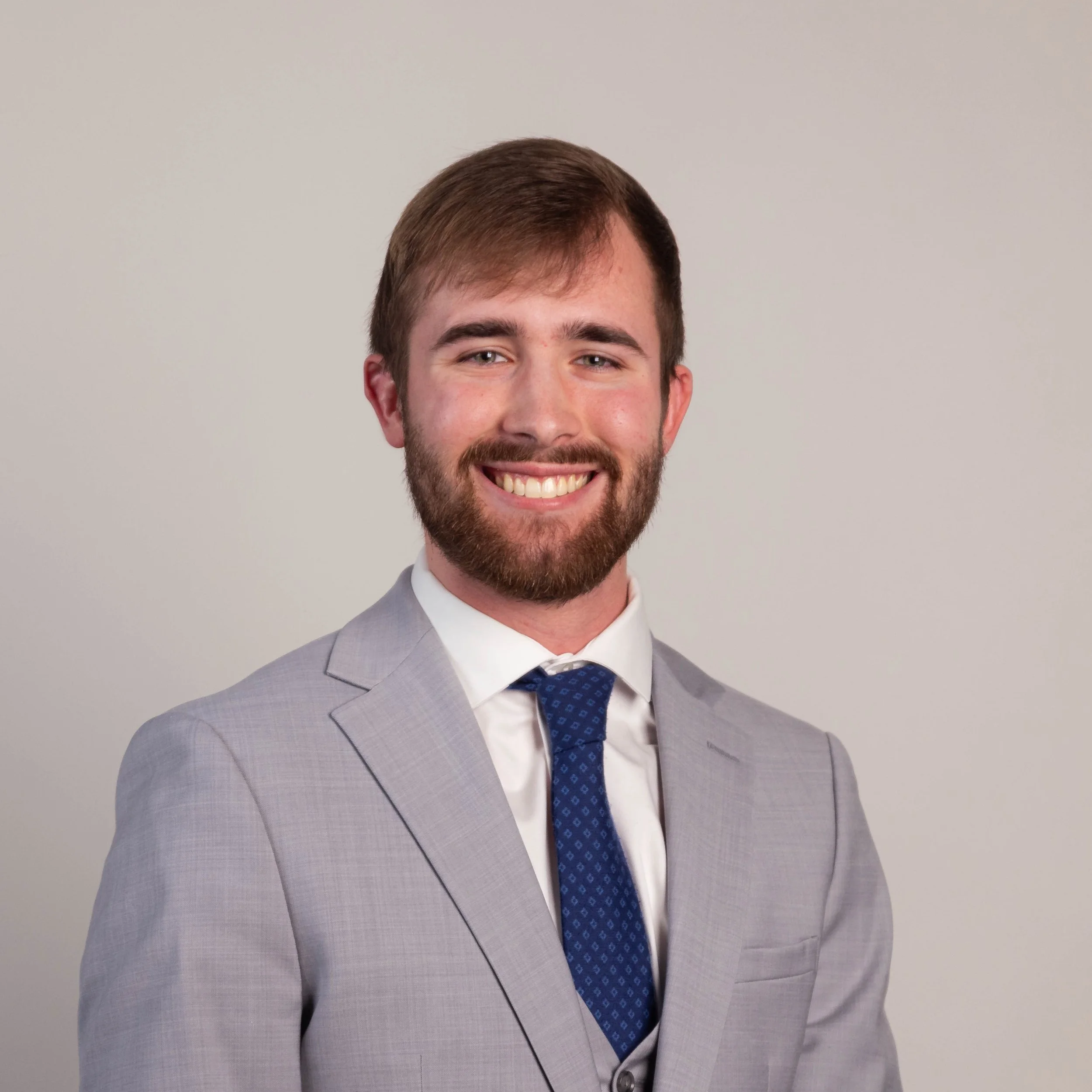 Photo of a young man with short brown hair and a beard, wearing a light gray suit, white shirt, and dark blue tie, smiling against a plain light gray background.