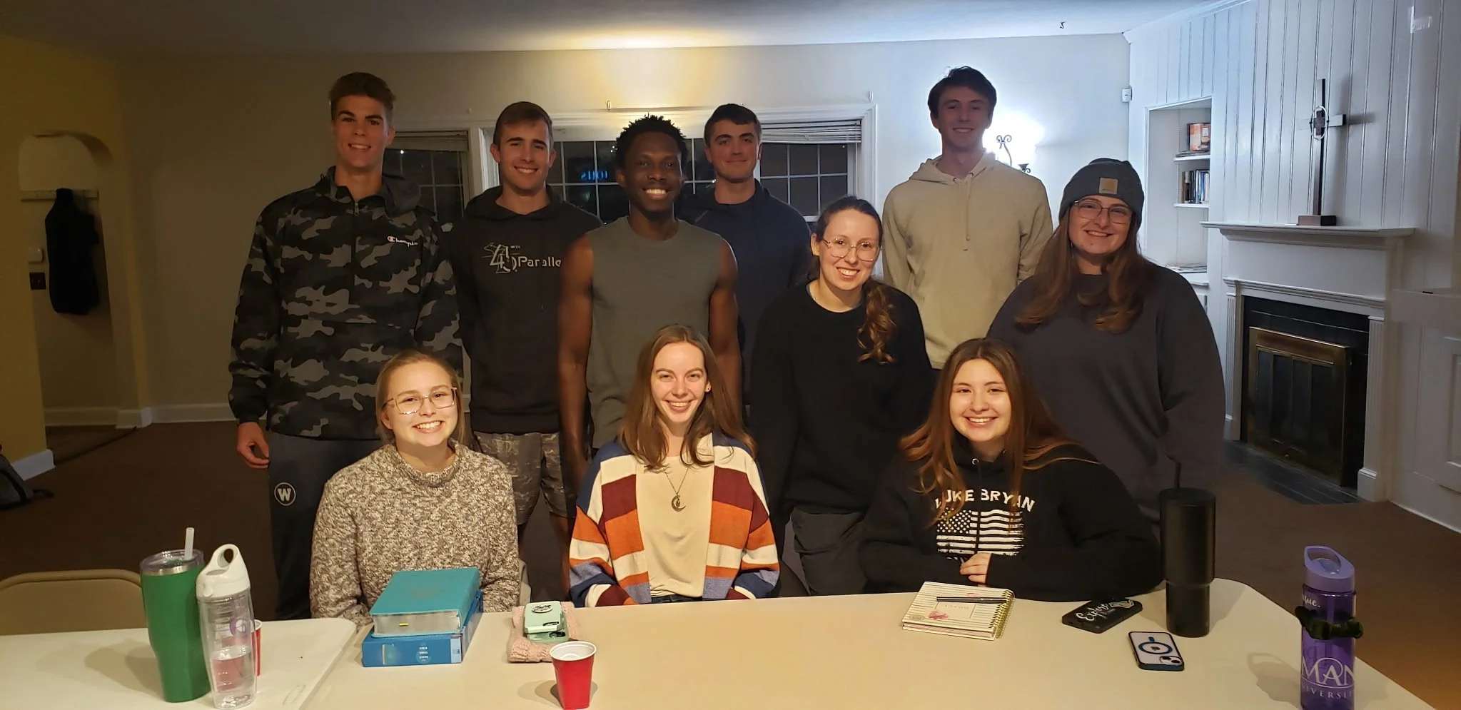 A group of ten young adults, five males and five females, gathered indoors in a room with white walls and fireplace, smiling at the camera. They are seated and standing around a table with notebooks, drinks, and personal items.