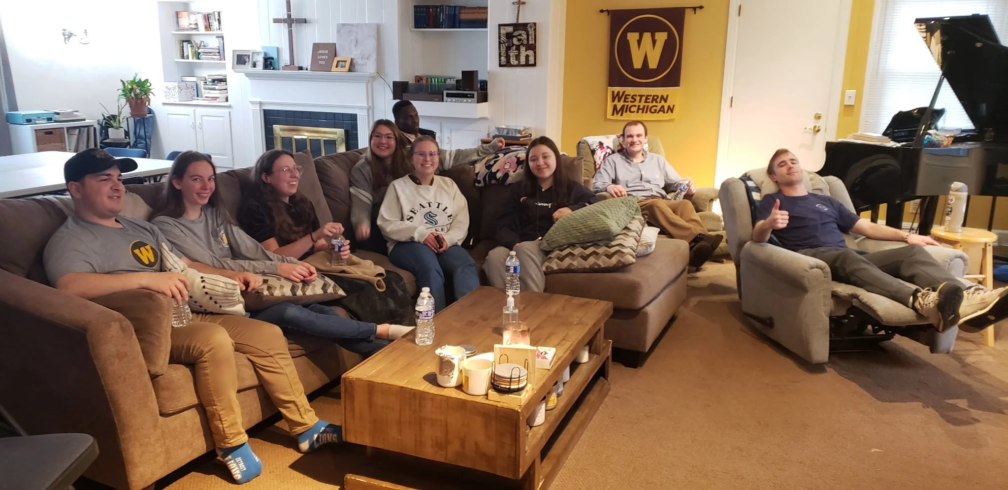 A group of people sitting on a brown and beige couch in a living room, smiling and looking at the camera. The room has white walls, a fireplace with a black and gold screen, and a yellow wall with a Western Michigan University banner. A black grand piano is in the corner, and a wooden coffee table with candles and cups is in front of the couch.
