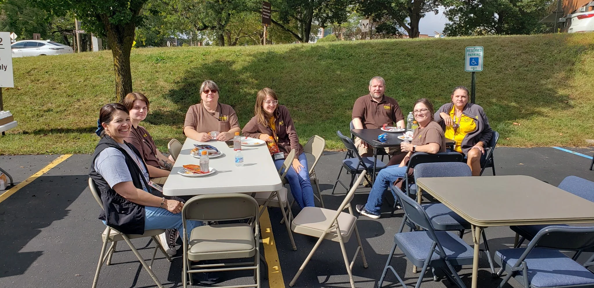 Group of seven people from the Western Michigan University staff dinner, sitting at outdoor tables, smiling and enjoying a meal, in a parking lot with trees and cars in the background.