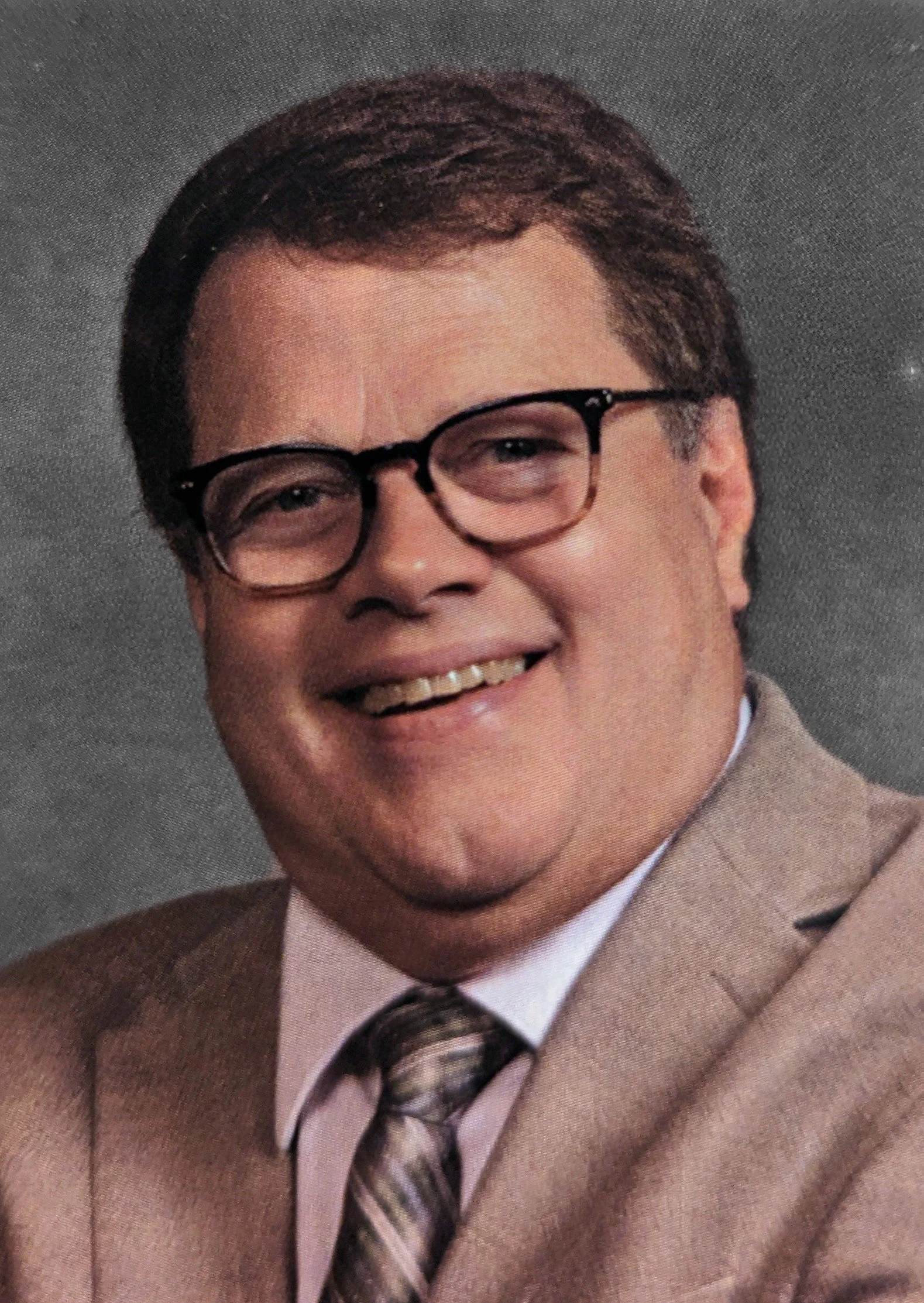 Close-up of a smiling man wearing glasses, a tan suit, a white shirt, and a patterned brown tie.
