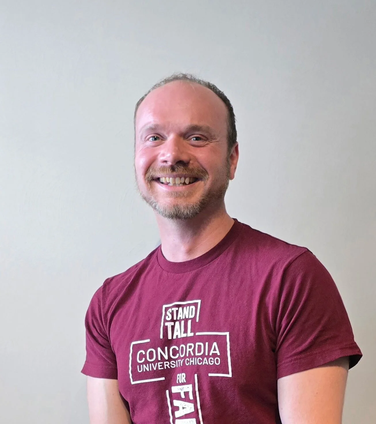 A smiling man with short hair, a beard, and a maroon Concordia University Chicago T-shirt standing in front of a plain light-colored wall.