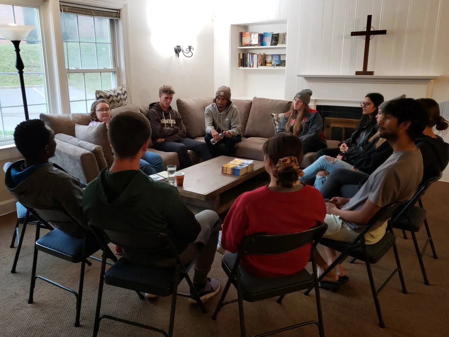 A group of diverse young people sitting in a circle in a cozy living room, engaged in a discussion, with a coffee table and bookshelf in the background.