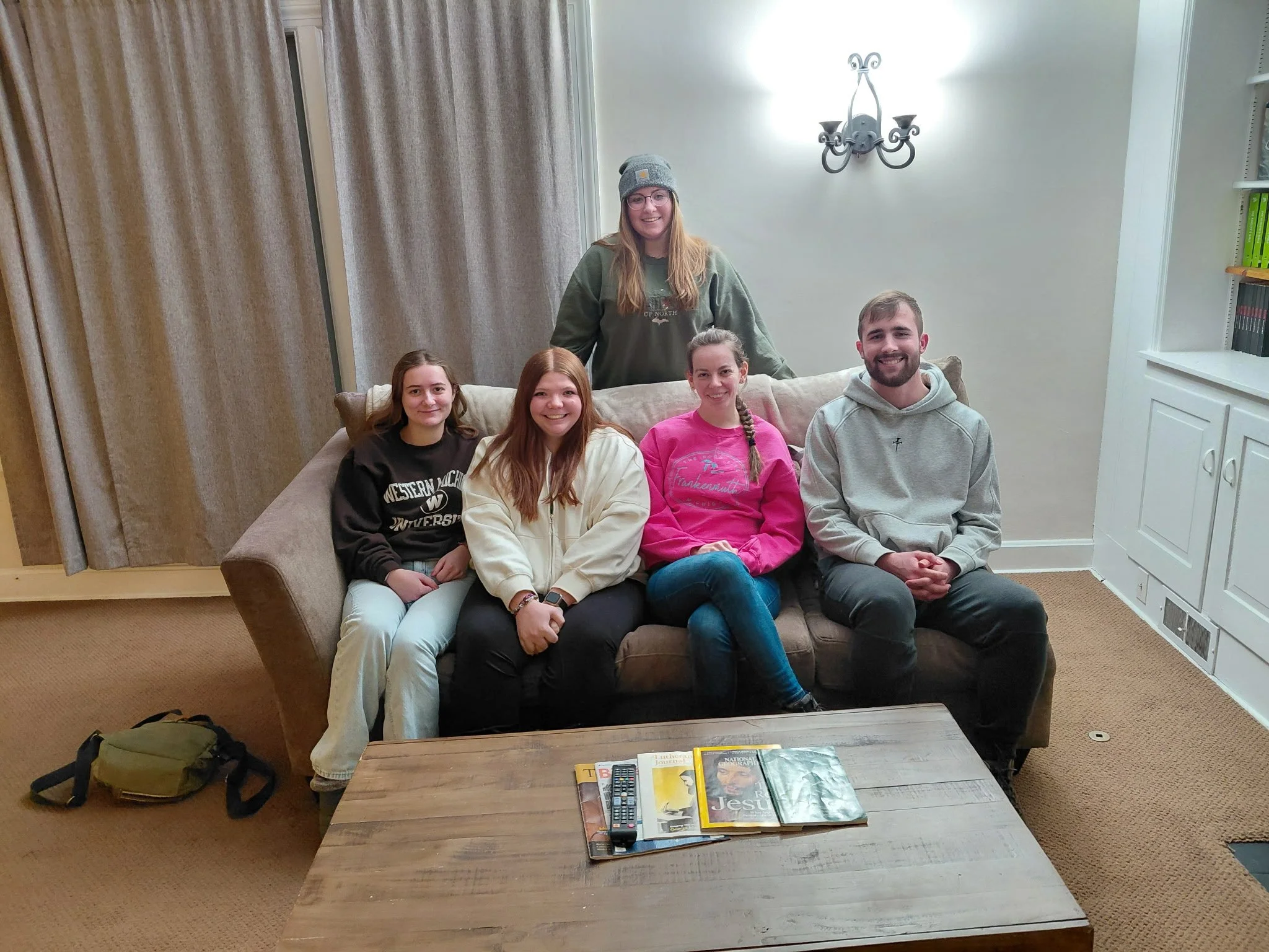 Group of six people sitting and standing on a sofa in a living room, with a coffee table in front of them and books and magazines on it.