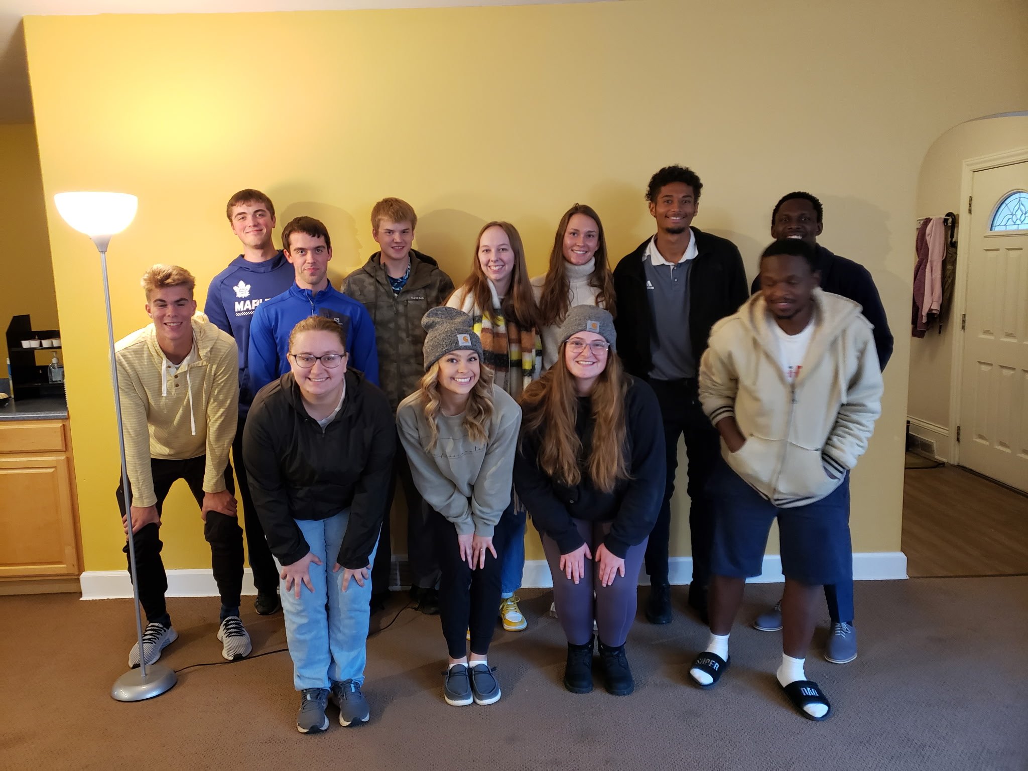 A group of fourteen young people posing indoors against a yellow wall, some standing and some kneeling, smiling at the camera.