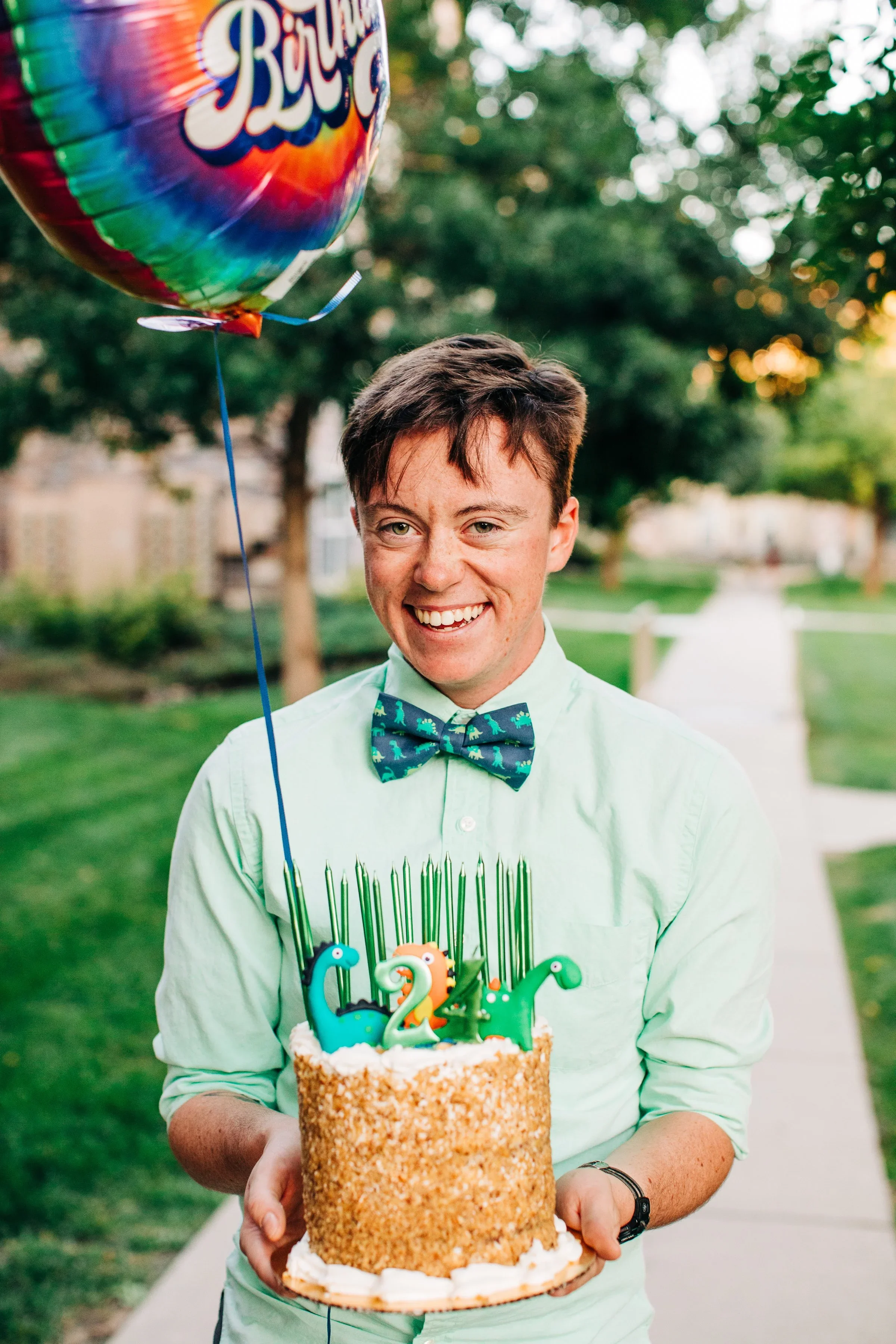Young man celebrating birthday outdoors with a cake decorated with colorful number candles and holding a rainbow birthday balloon.