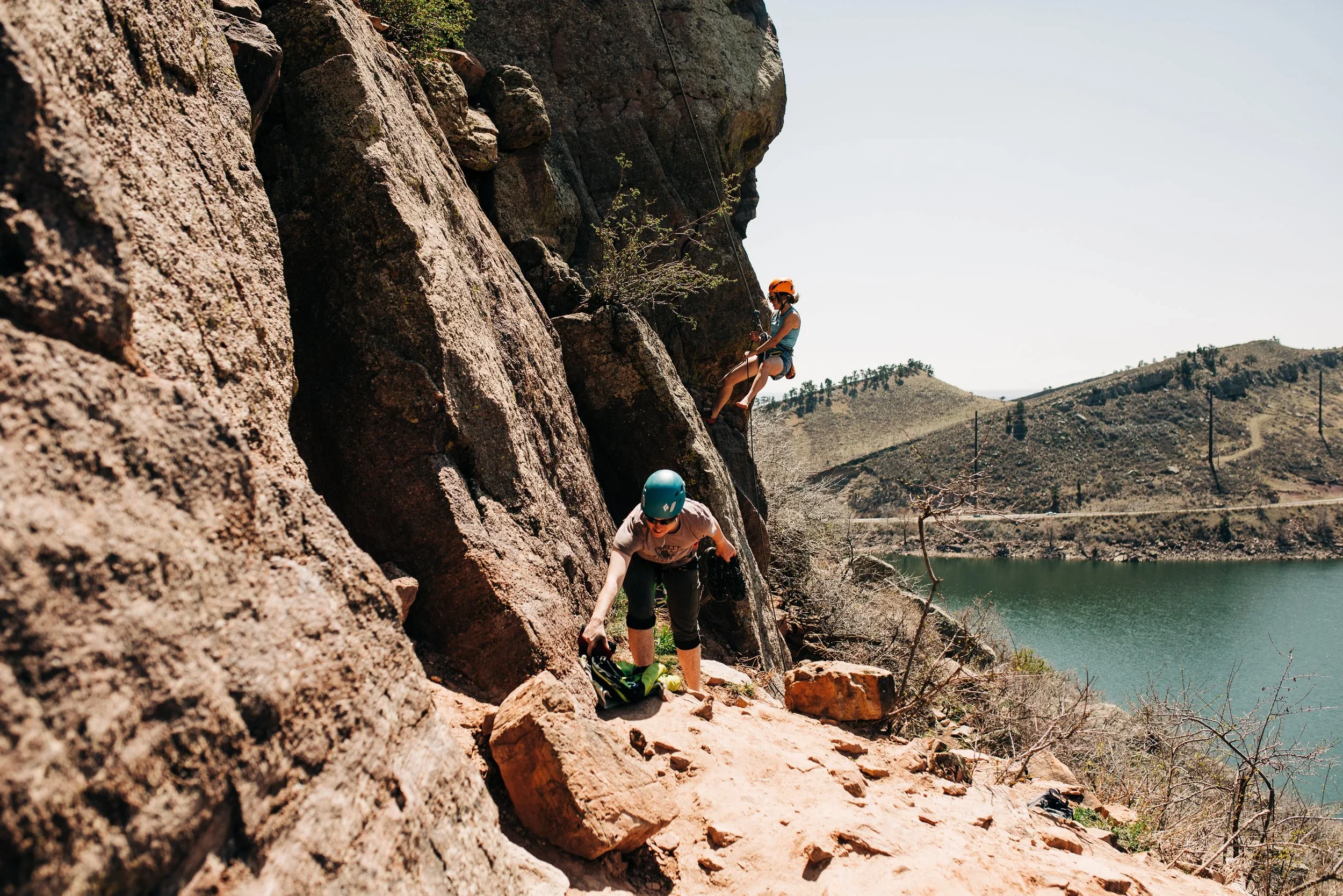 Two people rock climbing on a steep cliff by a body of water with dry, hilly terrain in the background. They are climbing at Horsetooth in Fort Collins, Colorado.
