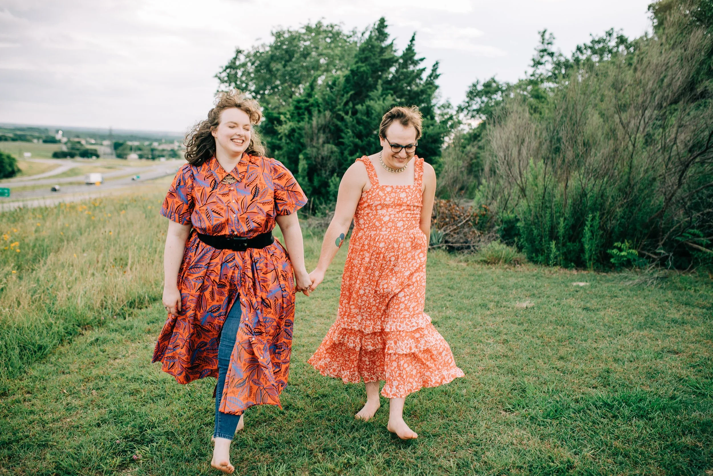 Two women holding hands and walking barefoot on a grassy field, with trees and a highway in the background, smiling and enjoying outdoor time on a cloudy day.