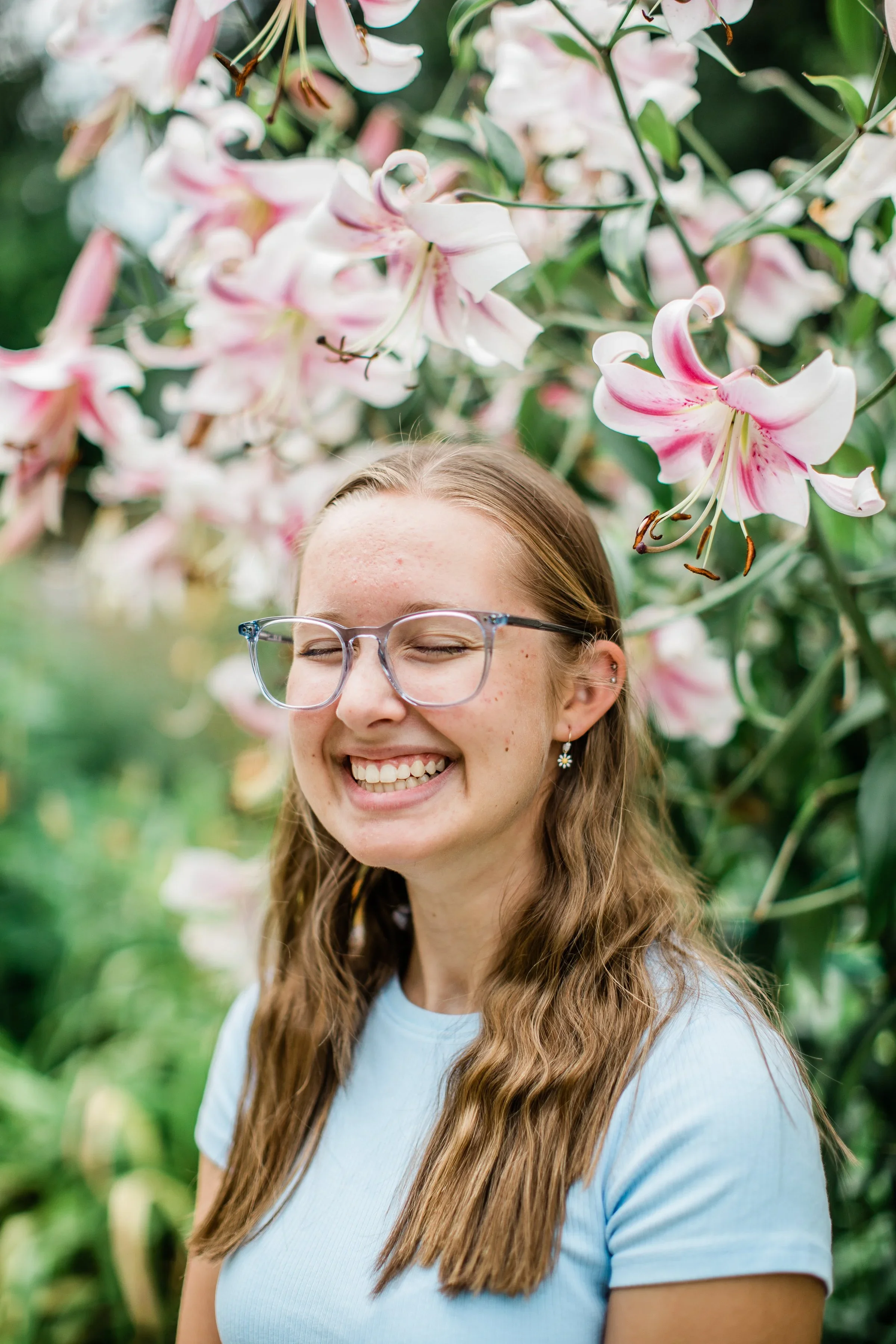 A young woman with long, wavy light brown hair, wearing glasses, earrings, and a light blue shirt, smiles with eyes closed in front of blooming pink and white lilies in a garden, the CSU Trial Garden in Fort Collins, Colorado