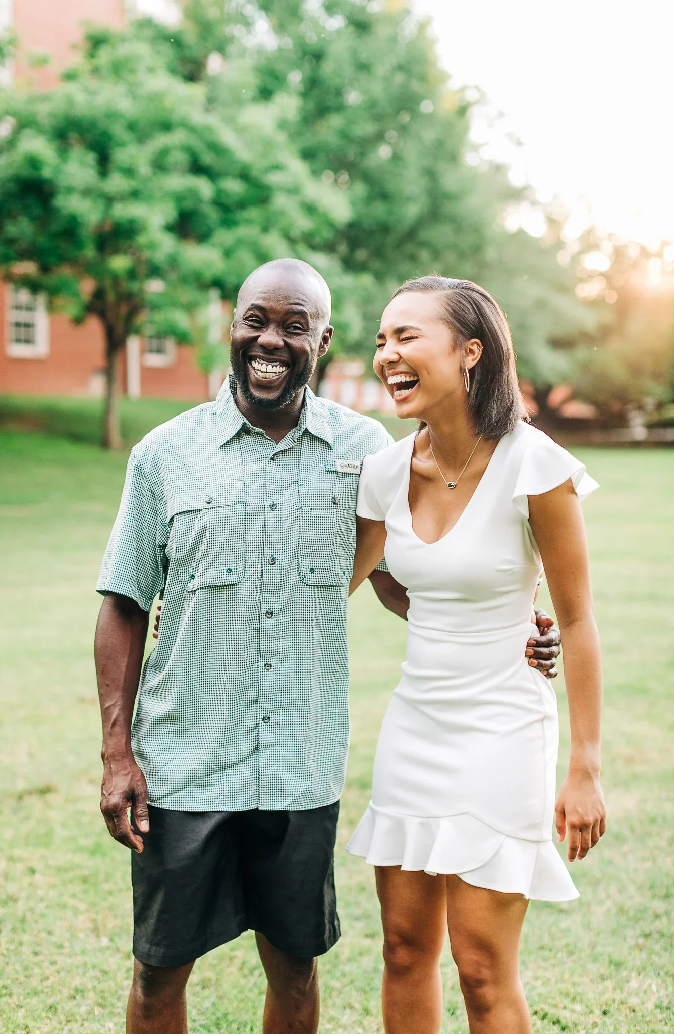 A man and a woman standing outdoors on a grassy area, smiling and laughing, with trees and a building in the background during sunset.