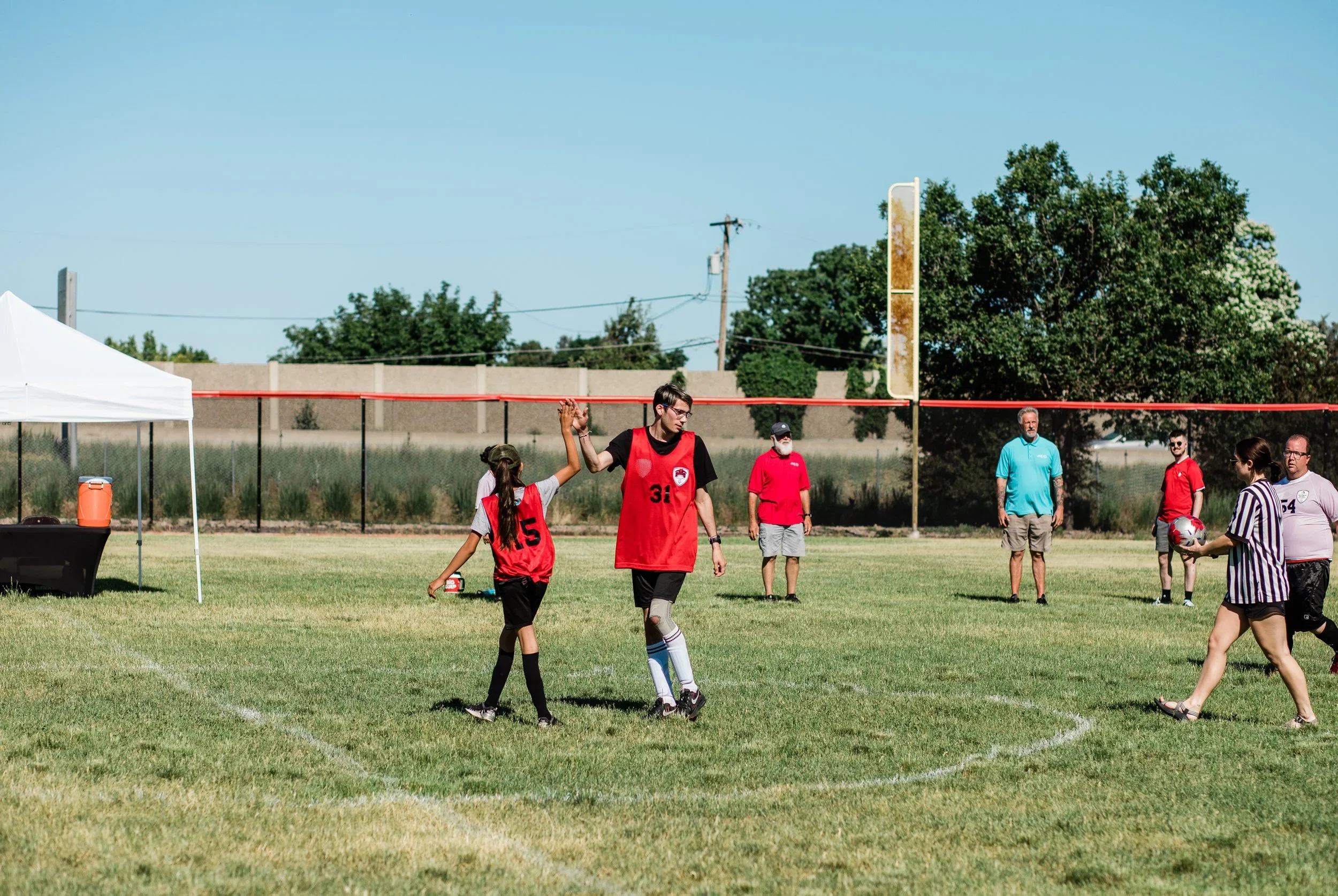 A youth soccer game at the Utah Special Olympics takes place on a grassy field with players, referees, and spectators, under a clear blue sky.