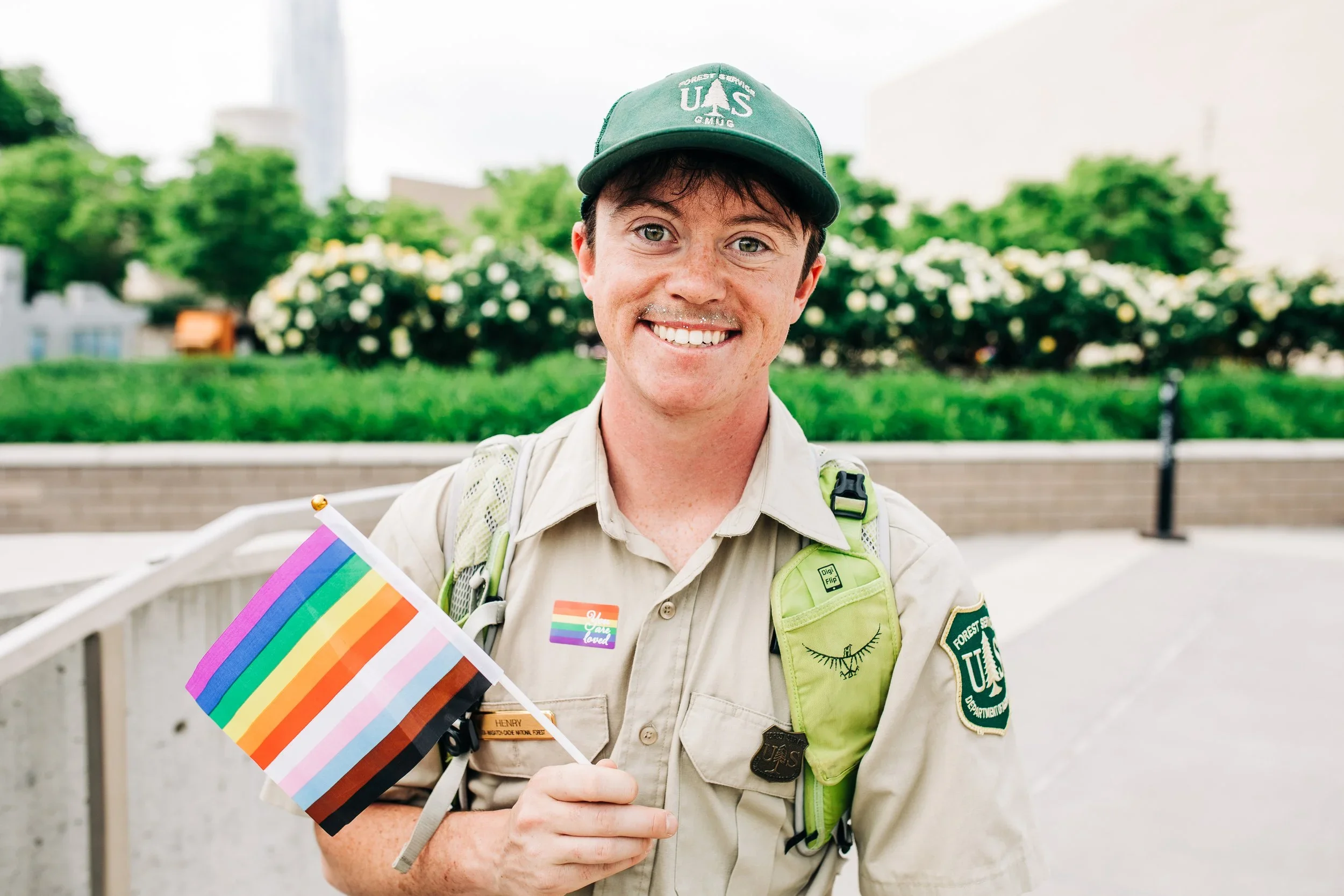 A young man dressed in a scout uniform holding a rainbow Pride flag at Salt Lake City Pride festival smiling with greenery and flowers in the background.