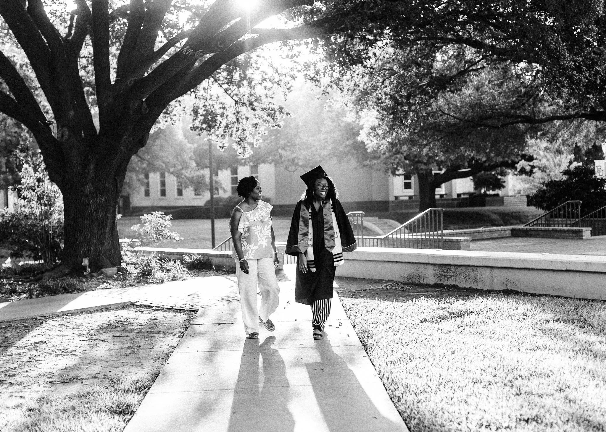A Black woman graduate in cap and gown walking with her mother on a college campus, beneath large trees on a sunny day.