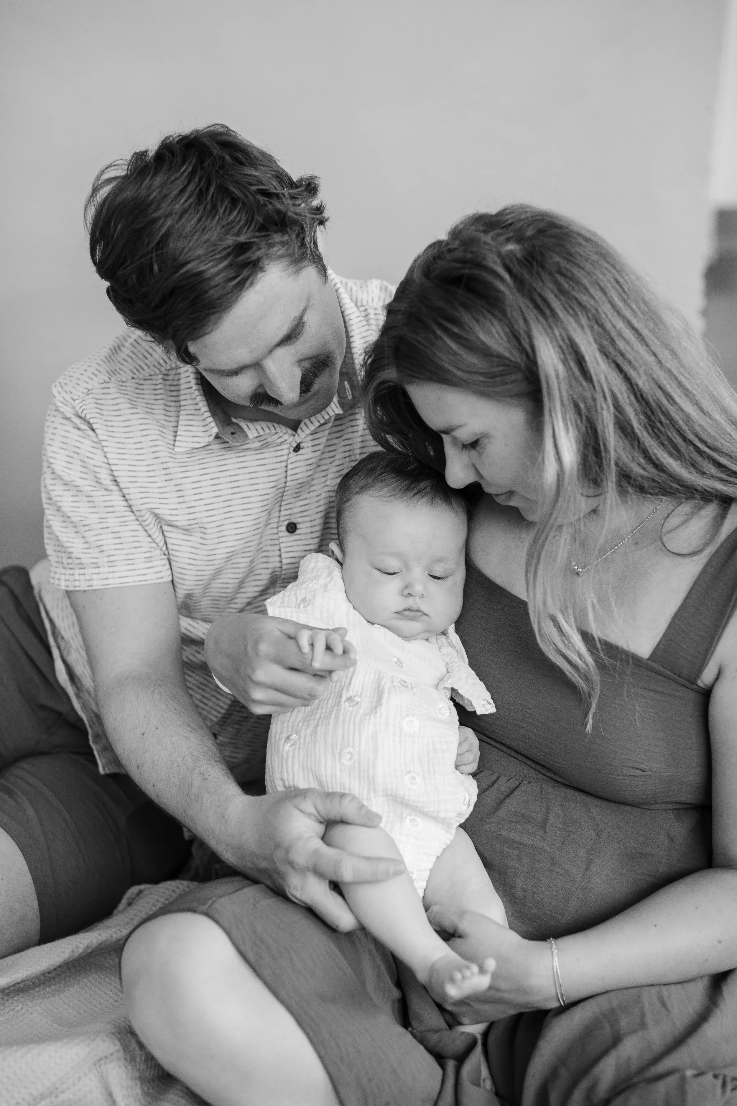 Black and white photo of a family with a baby taken in a Salt Lake City studio. The man and woman are leaning close to the baby, who is sitting on the woman's lap, with the man gently holding the baby's hand.