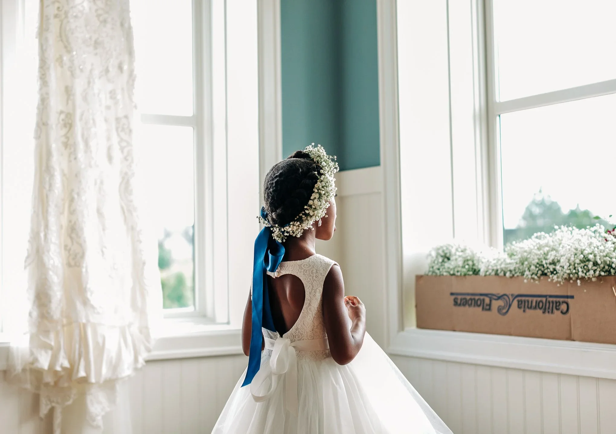 Young girl dressed in a white dress with a floral headband and blue ribbon, standing near a window with natural light.