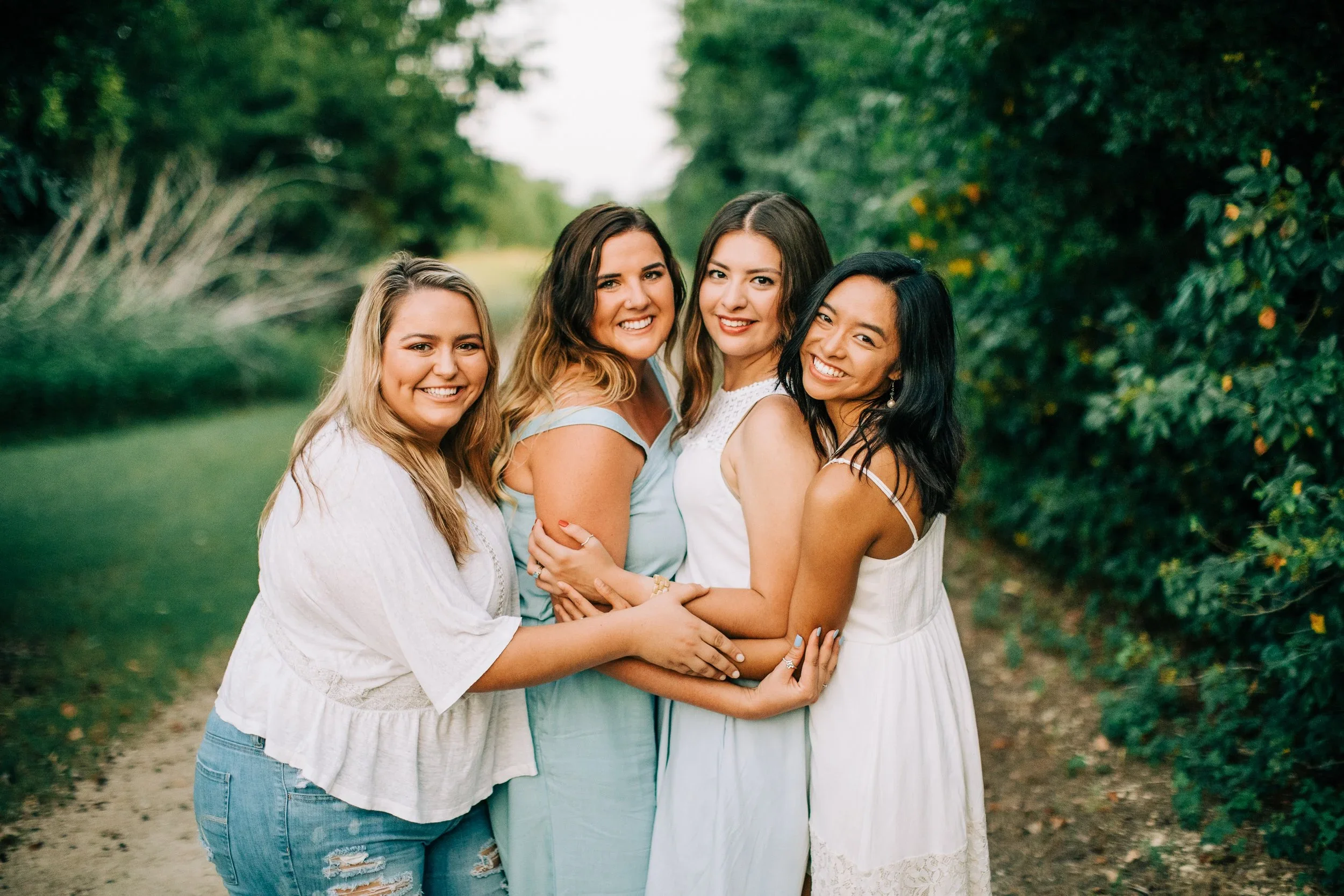 Five women standing close together outdoors on a path with greenery in the background, smiling at the camera.