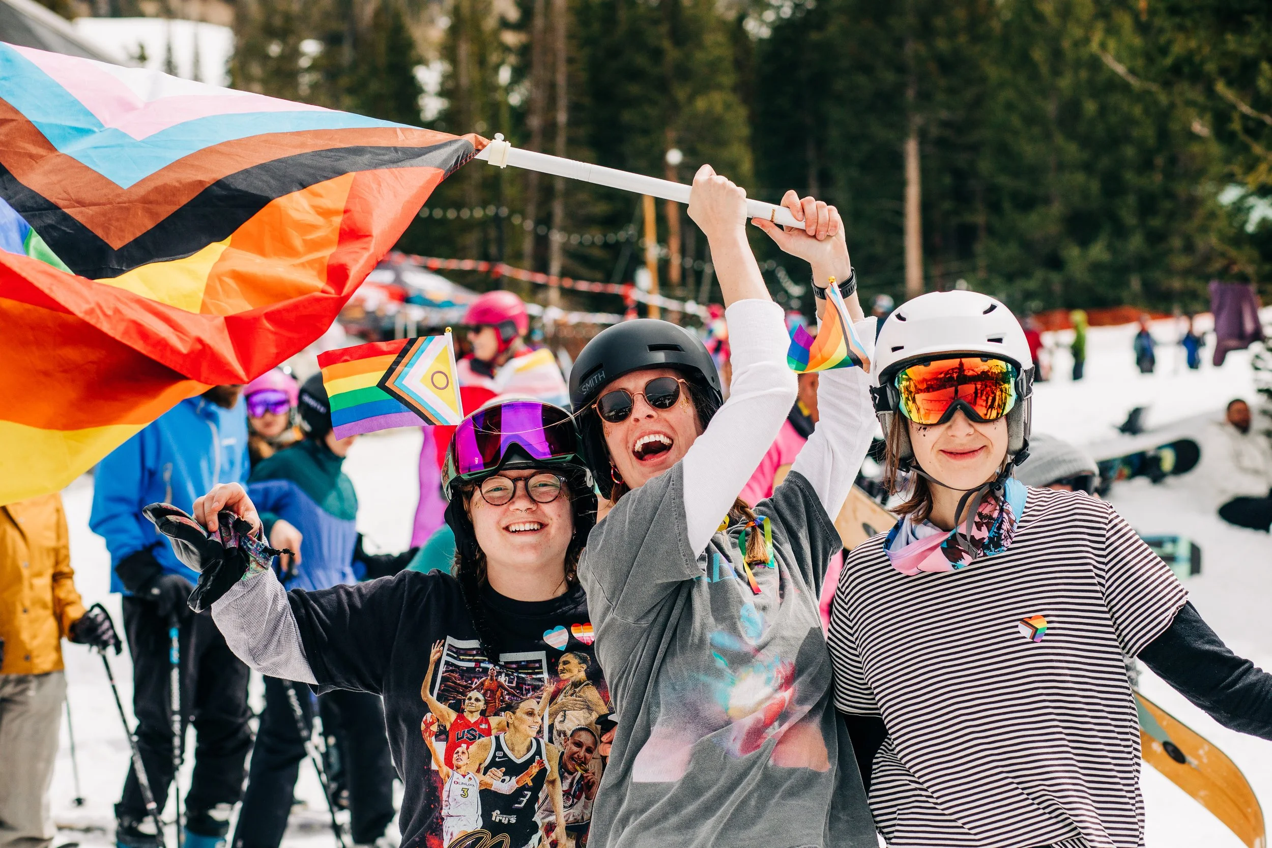Three women in ski helmets and goggles celebrating at a snowy outdoor ski resort, holding rainbow flags and waving a large rainbow flag, with other skiers and snowboarders in the background.