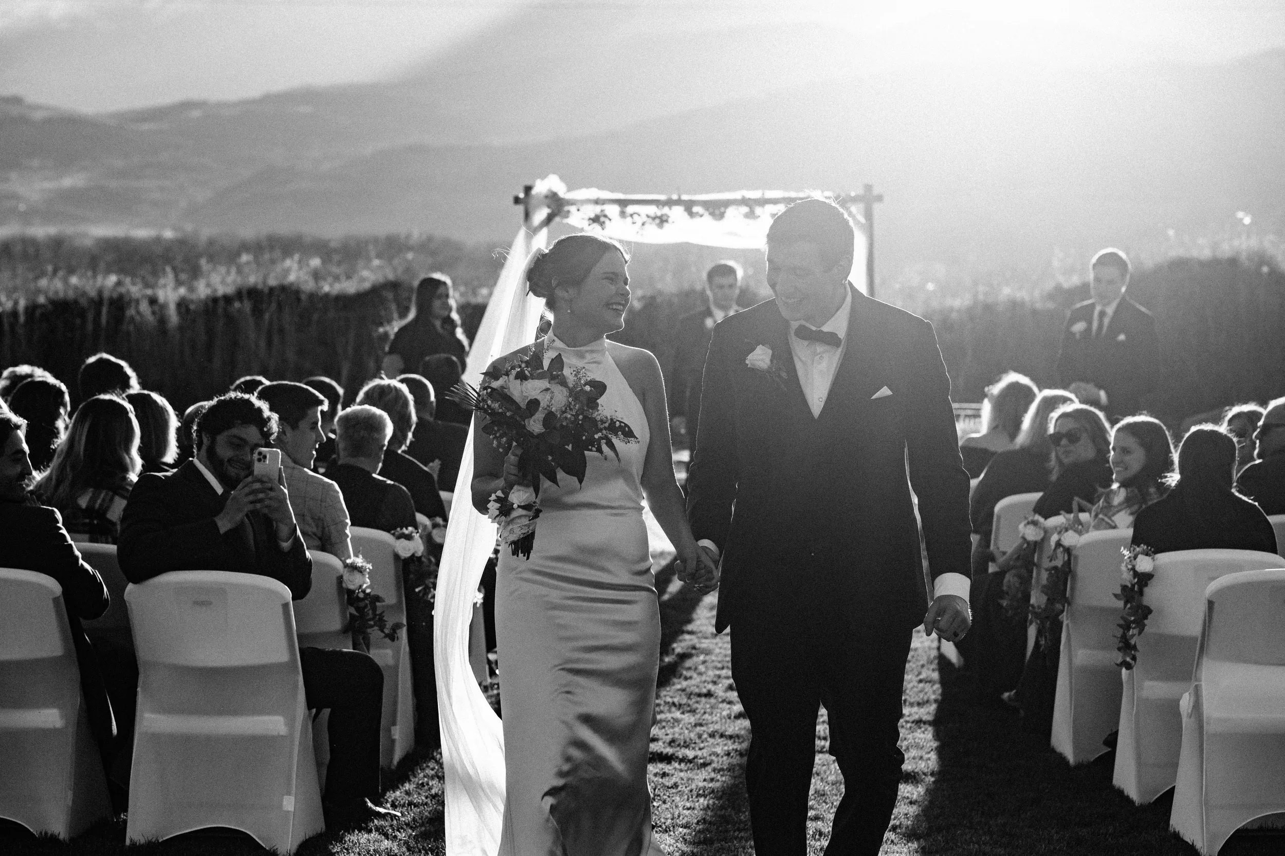 A bride and groom holding hands and smiling during their outdoor wedding ceremony at sunset in Morgan, UT, with guests seated in chairs on either side of the aisle.