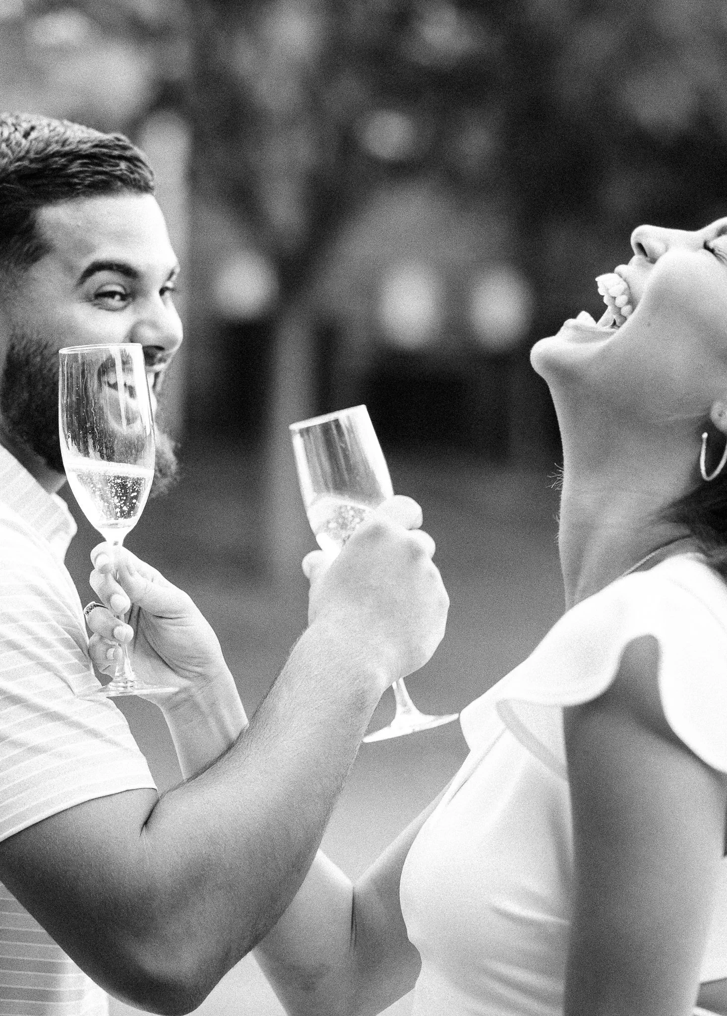A Black couple smiling and laughing while holding glasses of champagne, outdoors, just after their engagement photos.
