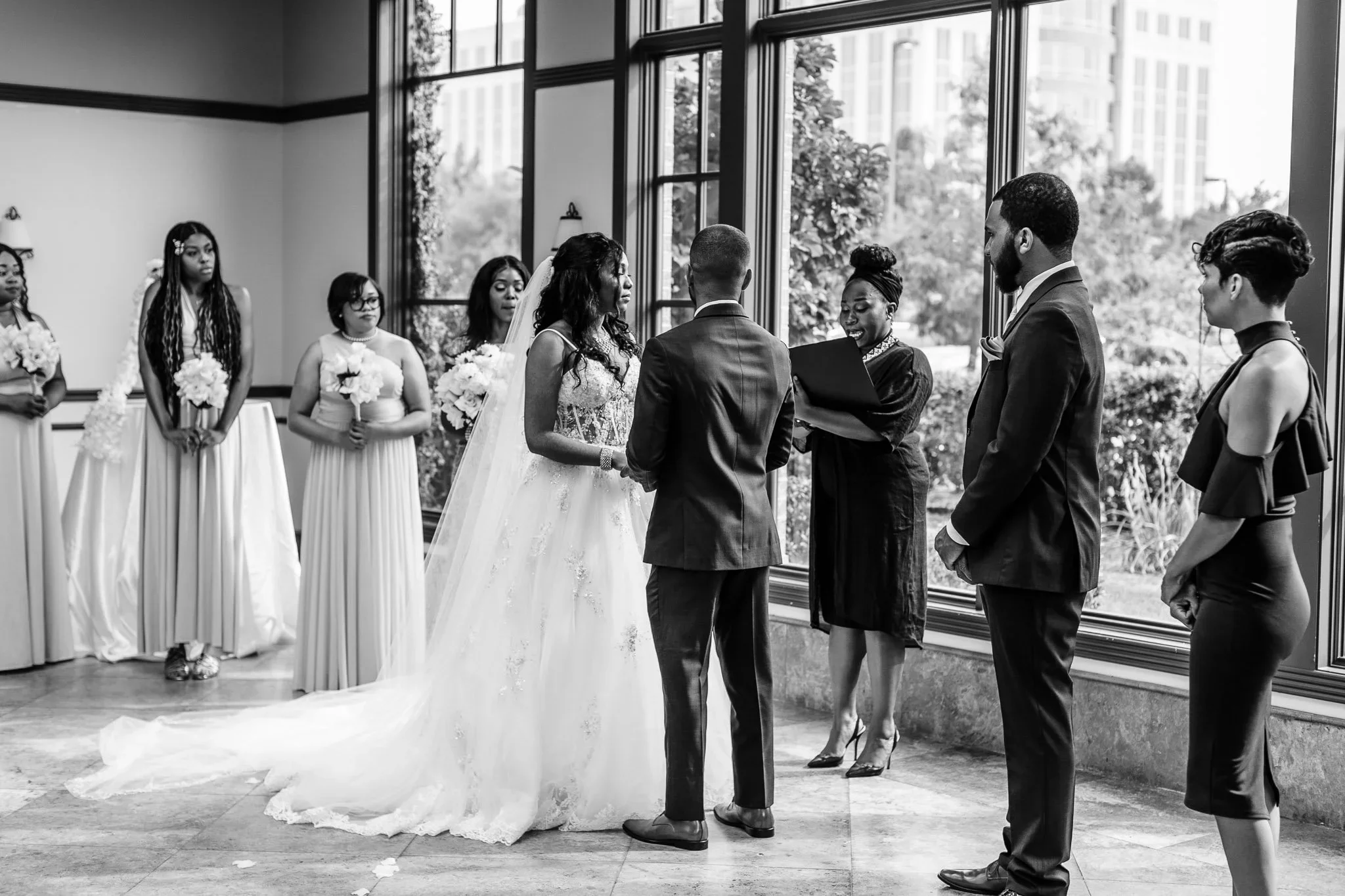 A black and white photo of a wedding ceremony with a Black bride and groom exchanging vows indoors near large windows, surrounded by BIPOC bridesmaids and groomsmen.