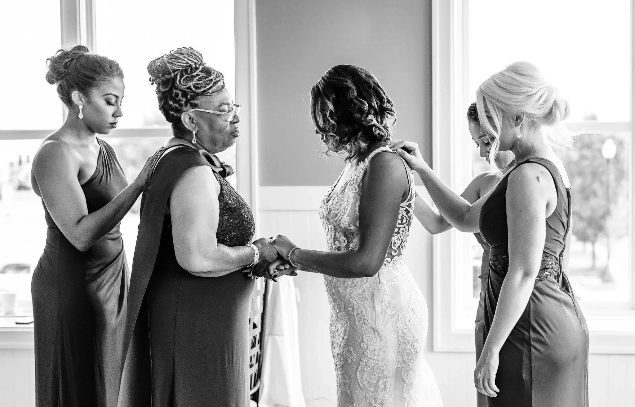 A Black bride in a wedding dress holding and praying with her mother, surrounded by her bridesmaids in dresses, inside a room with large windows.