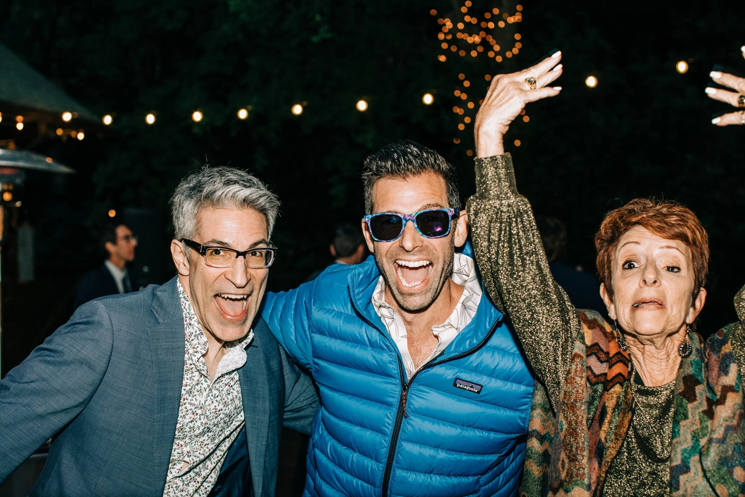 Three happy people celebrating at a party outdoors at night, smiling and dancing, with string lights overhead.