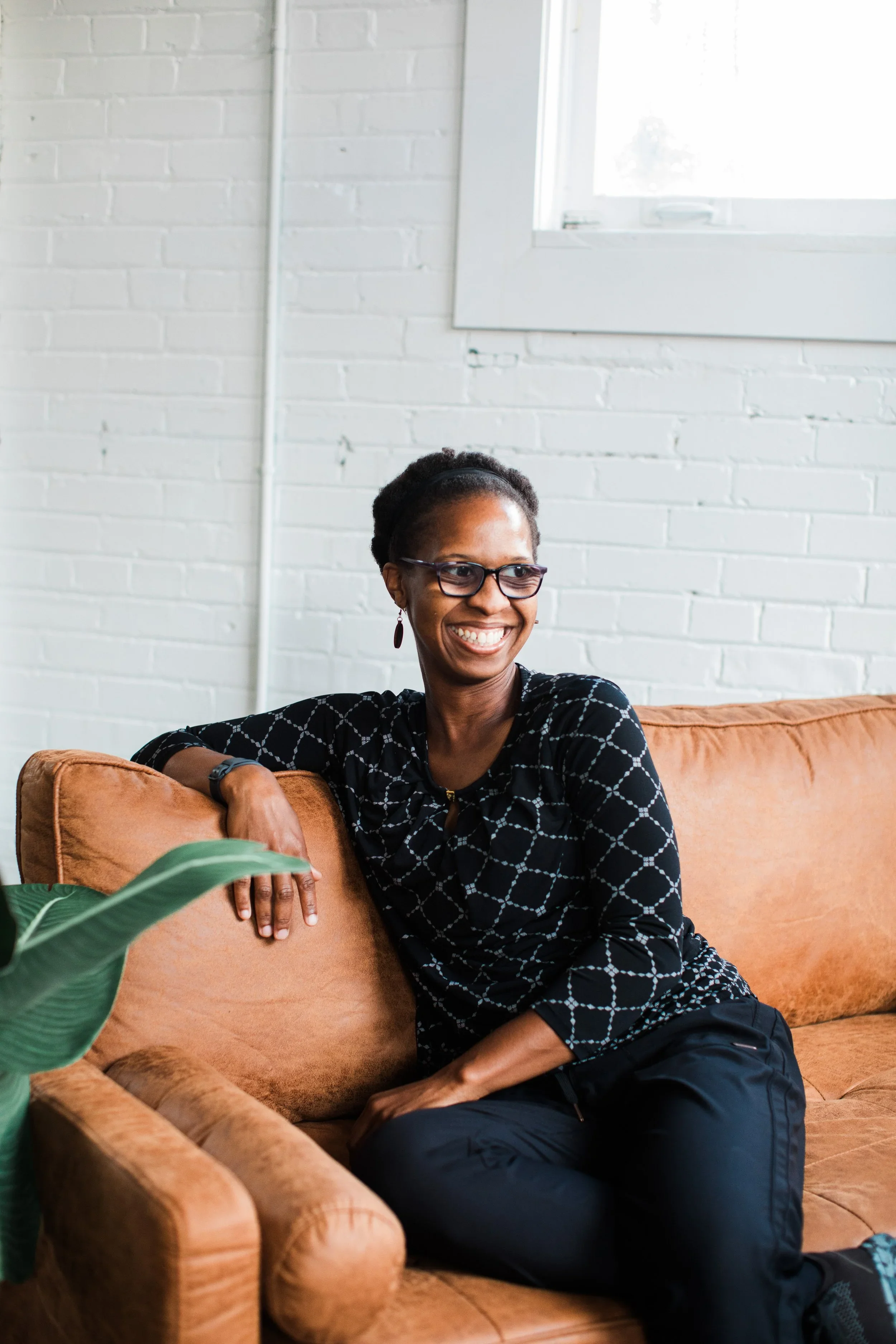 Smiling woman sitting on a tan sofa in a modern room with white brick walls and a large window, wearing glasses and black clothing.