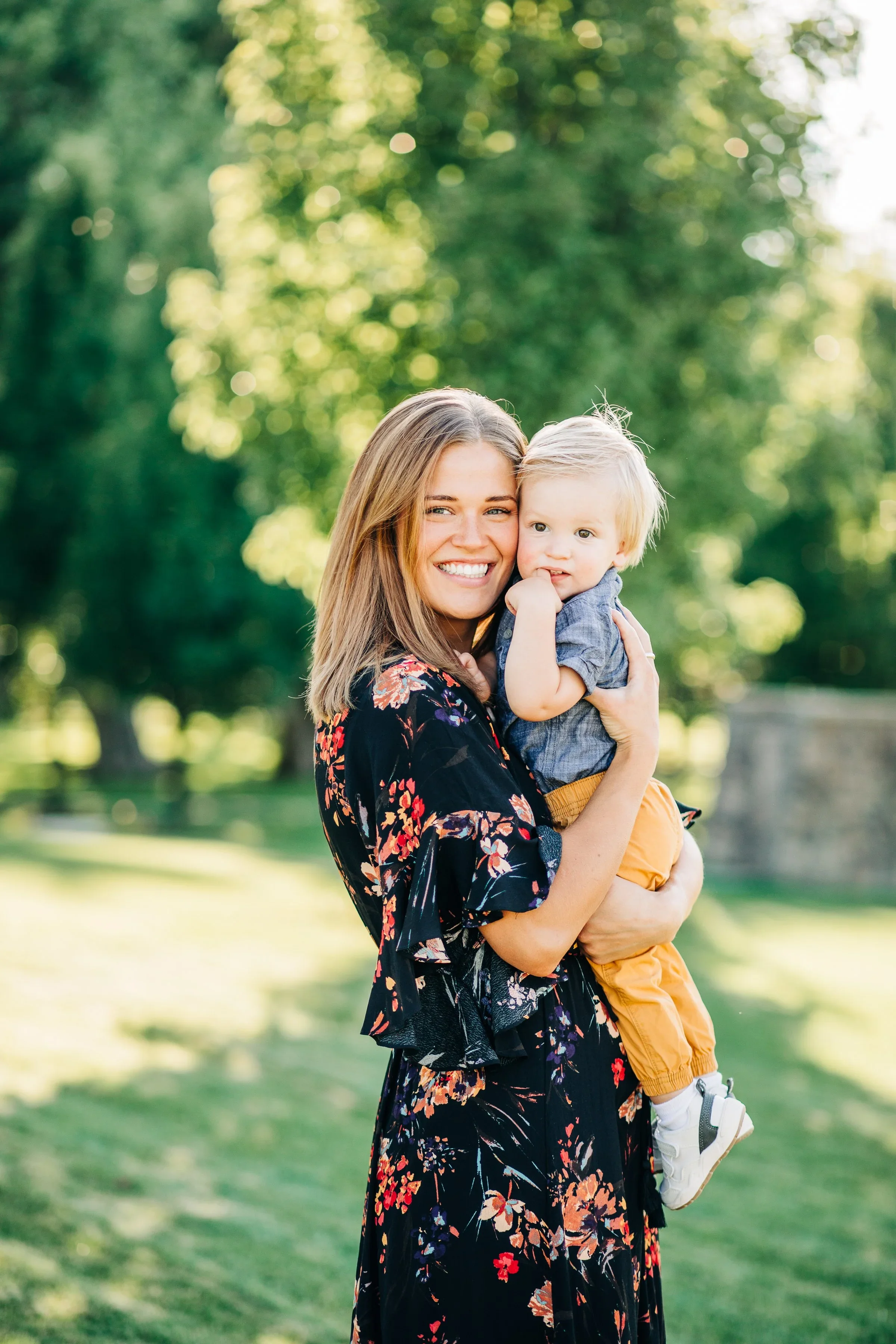 A mother holding a young child outdoors in a Fort Collins park with green trees and grass background.