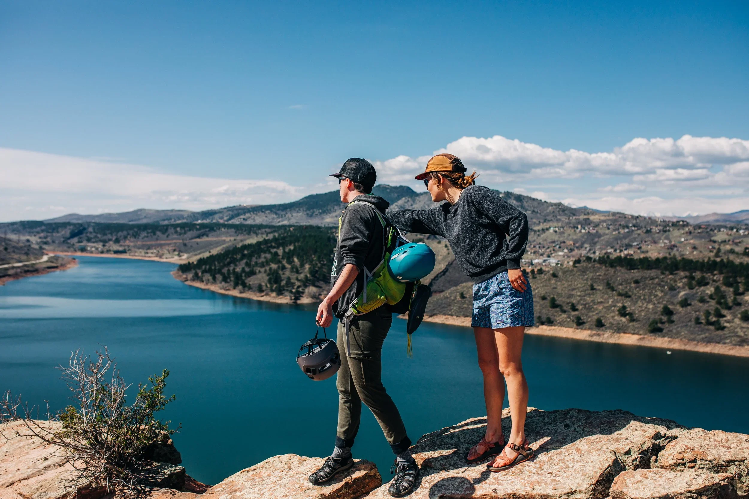 A man and woman standing on a rocky ledge overlooking a lake and mountains, with the woman touching the man's shoulder.