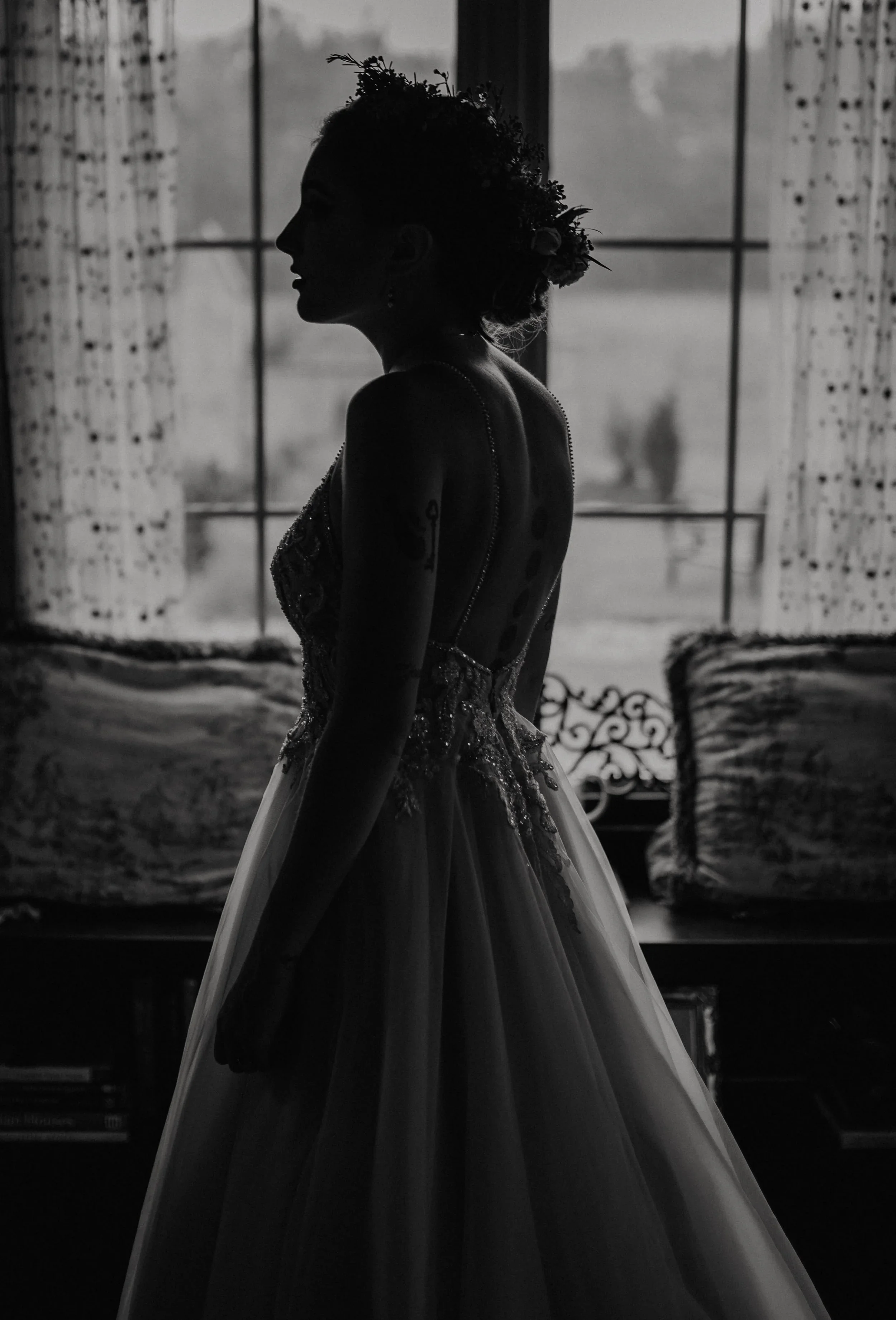 Silhouette of a woman in a wedding dress with a floral headpiece standing indoors near a window with curtains, in black and white.