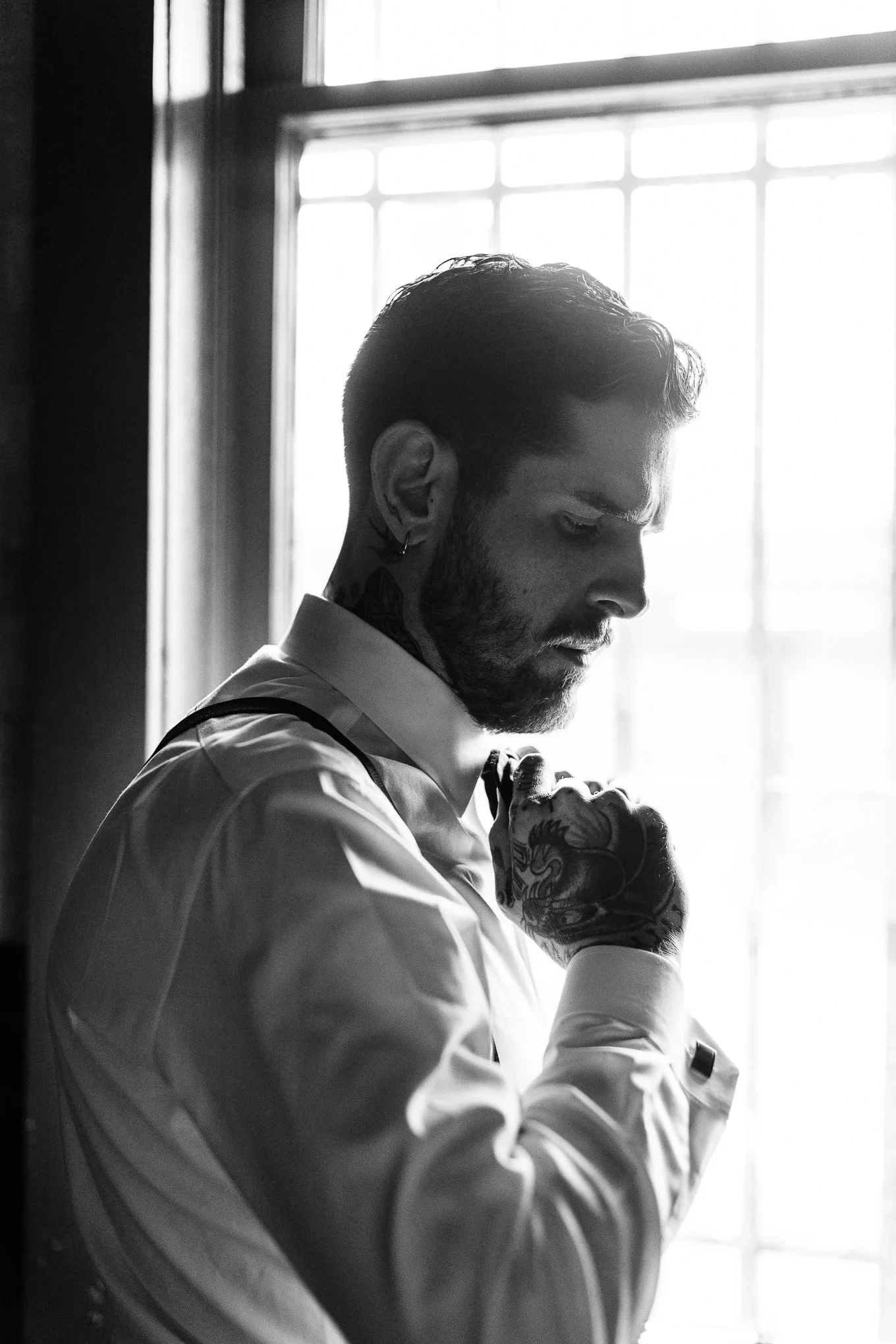 A man with tattoos, a beard, and a earring adjusts his tie while standing by a window, backlit with natural light.