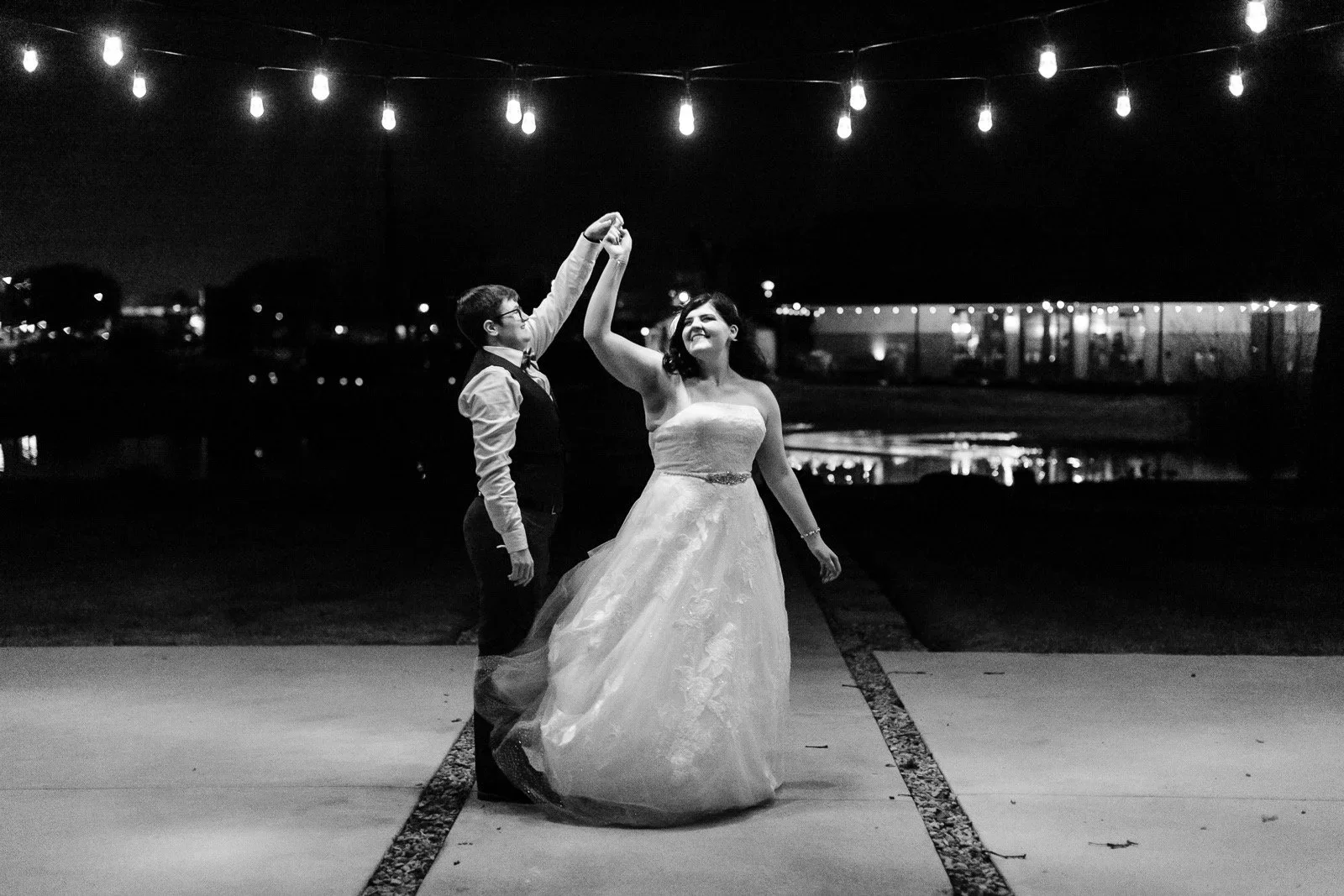 A queer couple, the woman in a wedding dress and the nonbinary person in a vest and shirt, dancing outdoors at night under string lights.