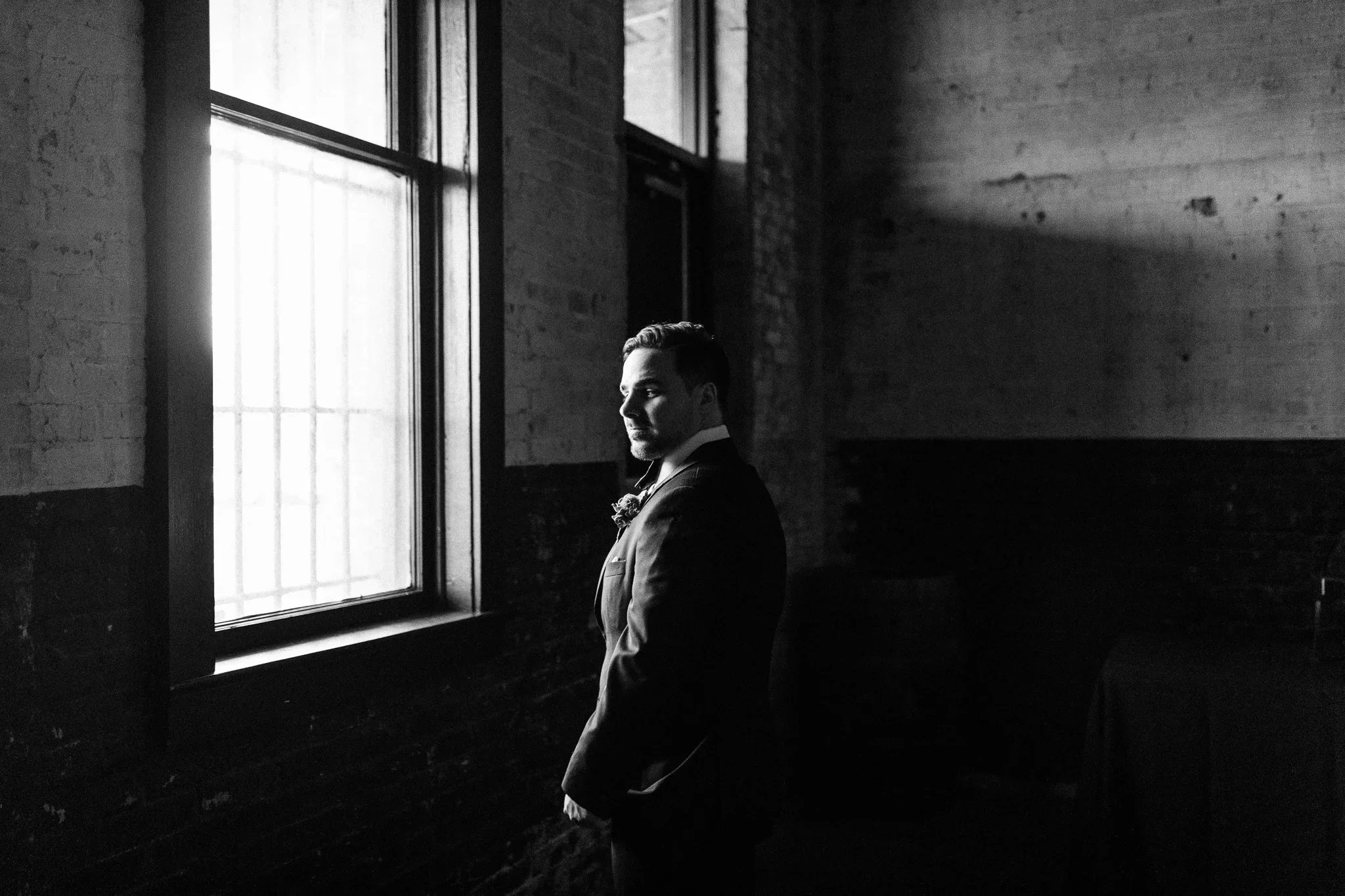 A man in a suit standing near a window in an industrial-style room with brick walls, black and white photo.