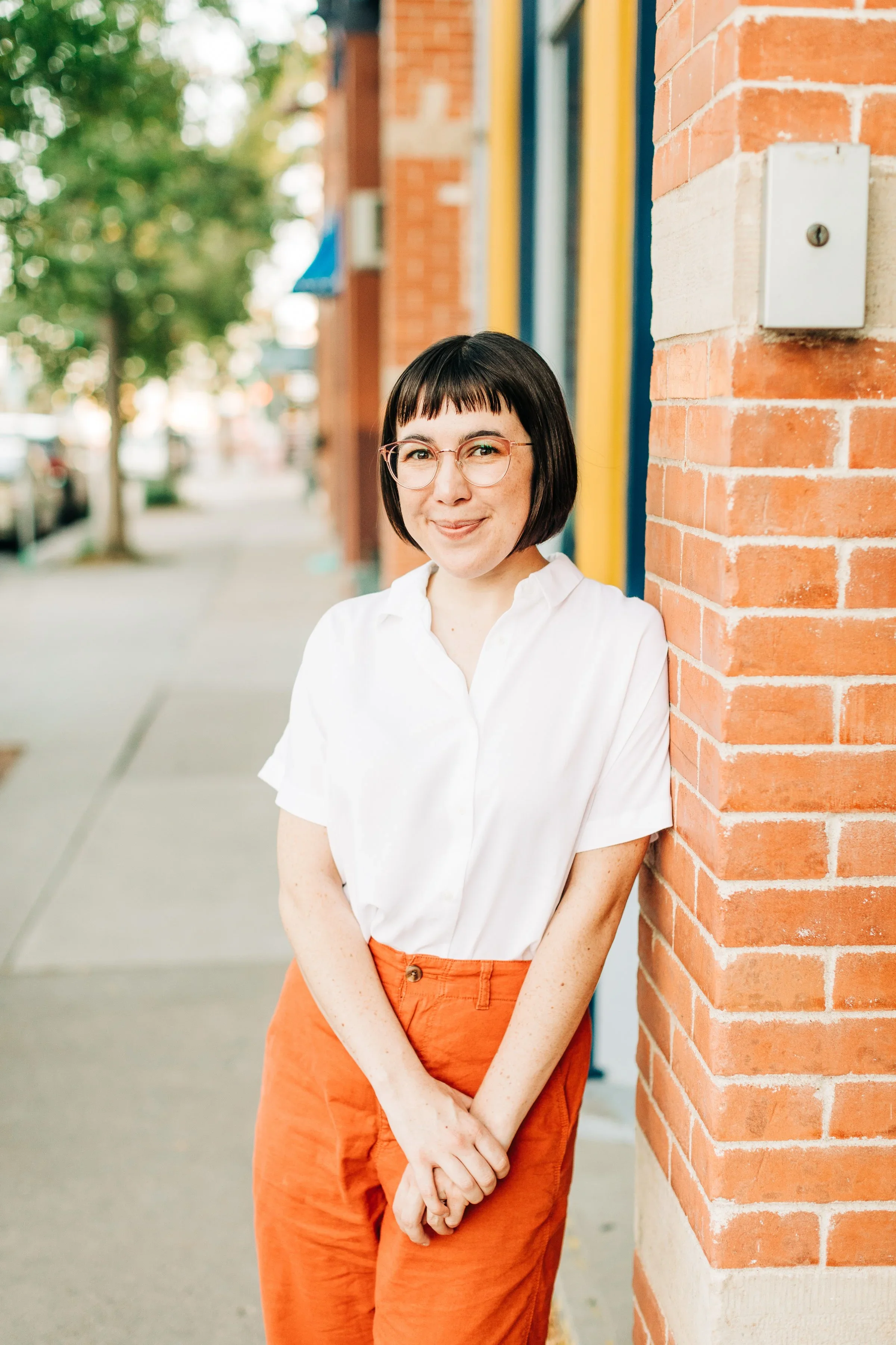 A woman with short black hair, glasses, wearing a white blouse and orange pants, leaning against a brick wall on a sidewalk, smiling at the camera.
